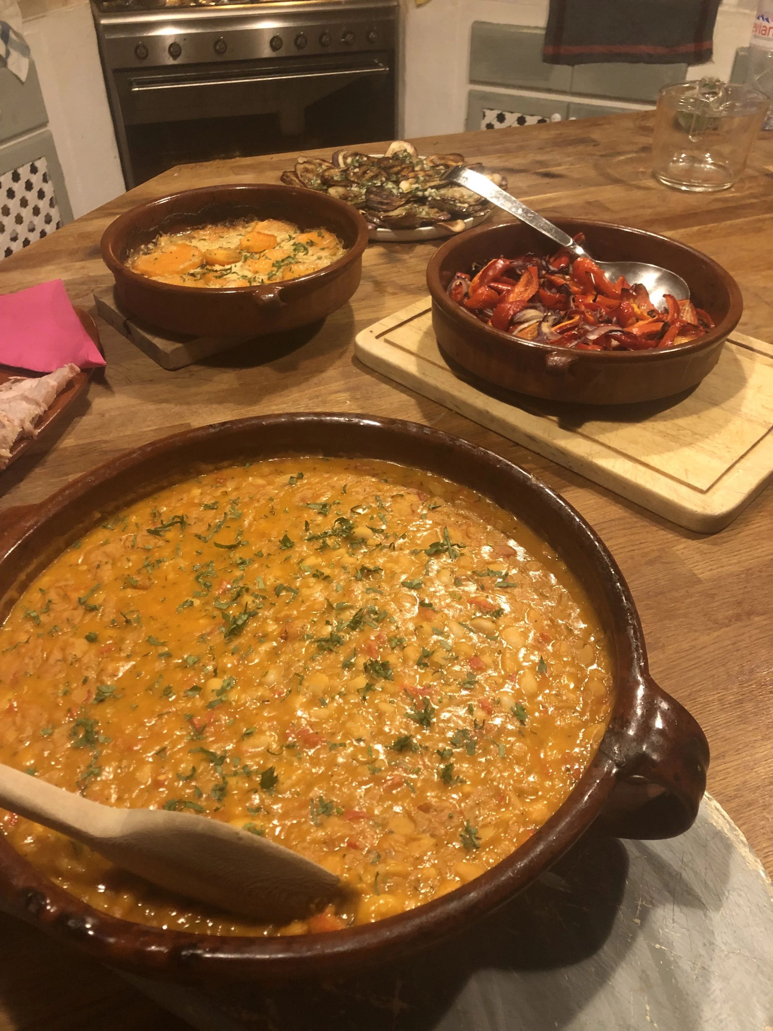 A kitchen table with several dishes of food, including a large dish of chili garnished with cilantro, a plate of roasted red peppers, and a bowl of roasted mushrooms. In the background, there is an oven and kitchen cabinets.