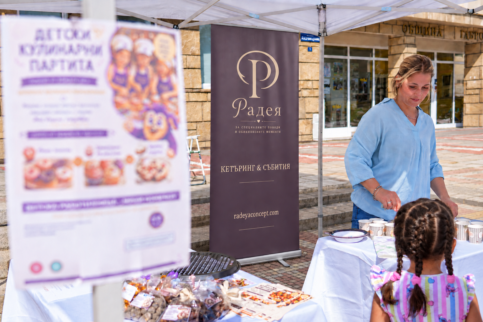 A woman at an outdoor booth serving at a table covered with cups and wrapped candies, with a young girl with braided hair facing the woman. Behind them is a banner that reads 'Радея' and a blurred poster in Bulgarian.