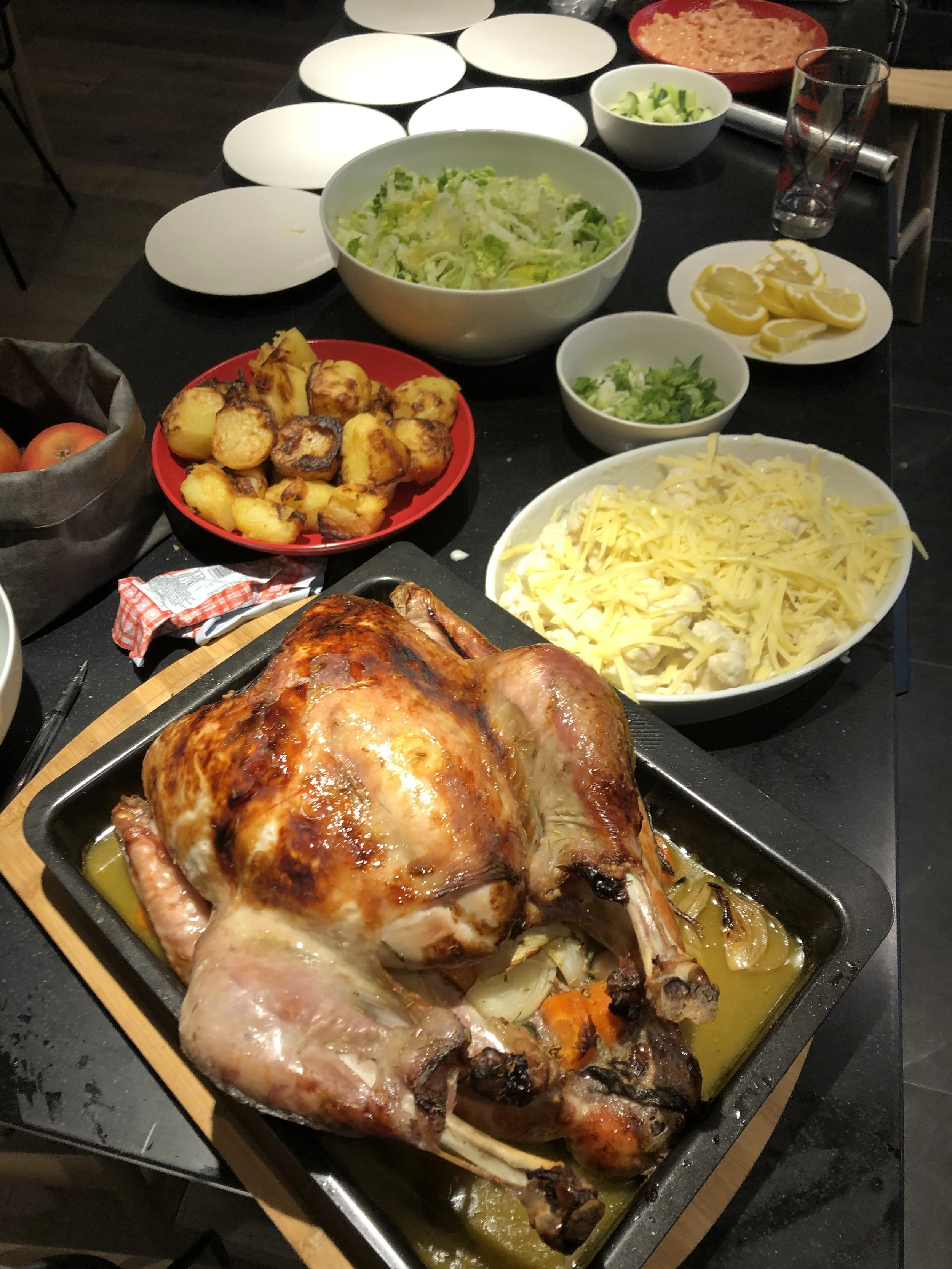 A roasted turkey on a black baking sheet with vegetables, surrounded by bowls of salad, lemon wedges, and various side dishes on a black table.