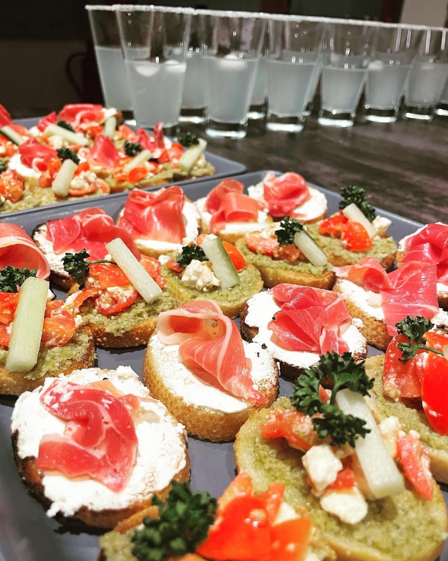 Assorted mini toasts with toppings including cream, smoked salmon, cherry tomatoes, herbs, and vegetables, arranged on a platter with glasses of water in the background.