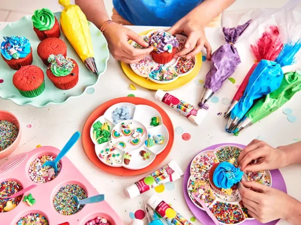 People decorating cupcakes with colorful frosting, sprinkles, and icing tubes, surrounded by various baking supplies and colorful cupcake liners.