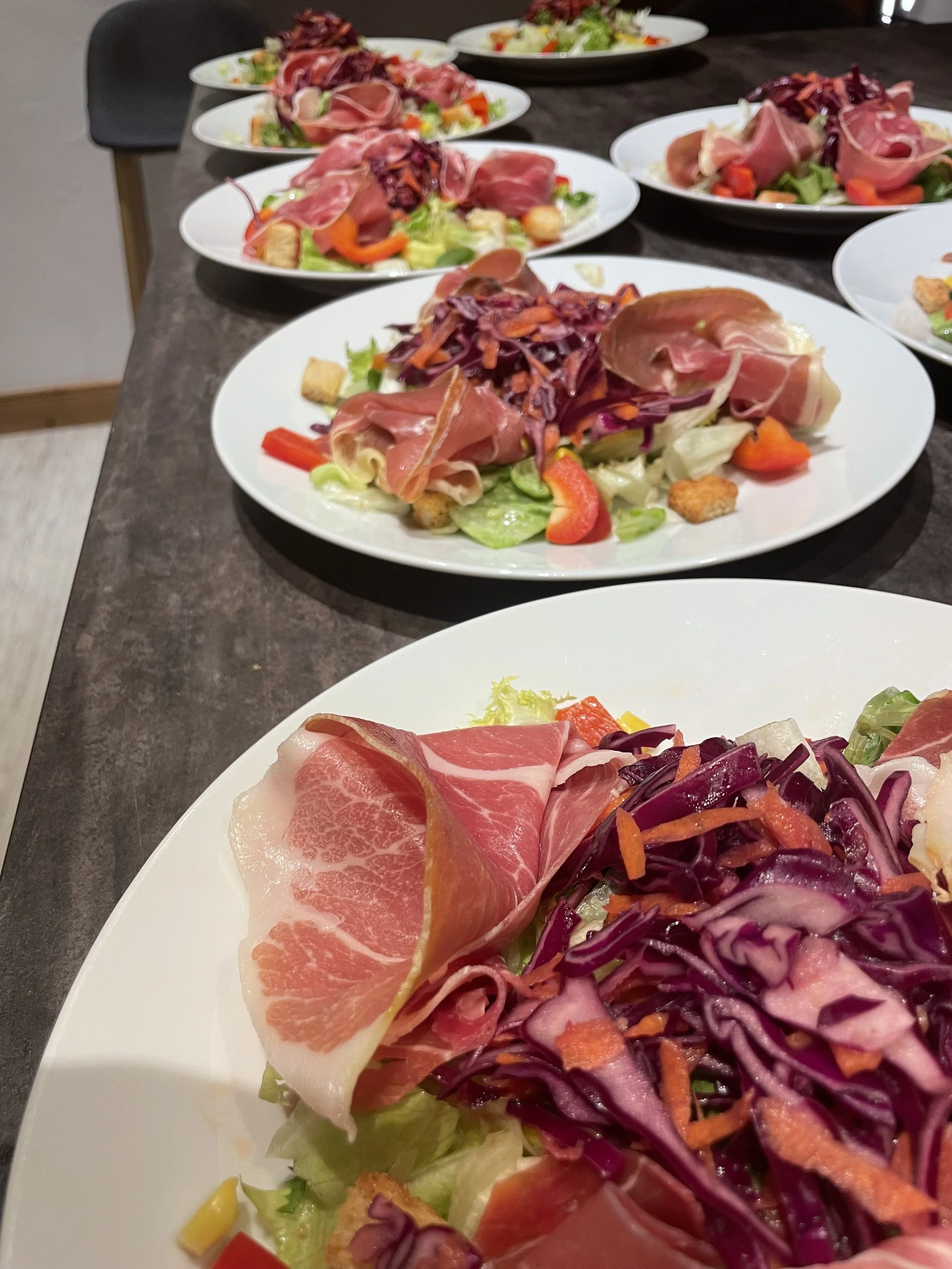A plate of assorted salads with sliced prosciutto, shredded purple and orange carrots, chopped lettuce, and croutons.