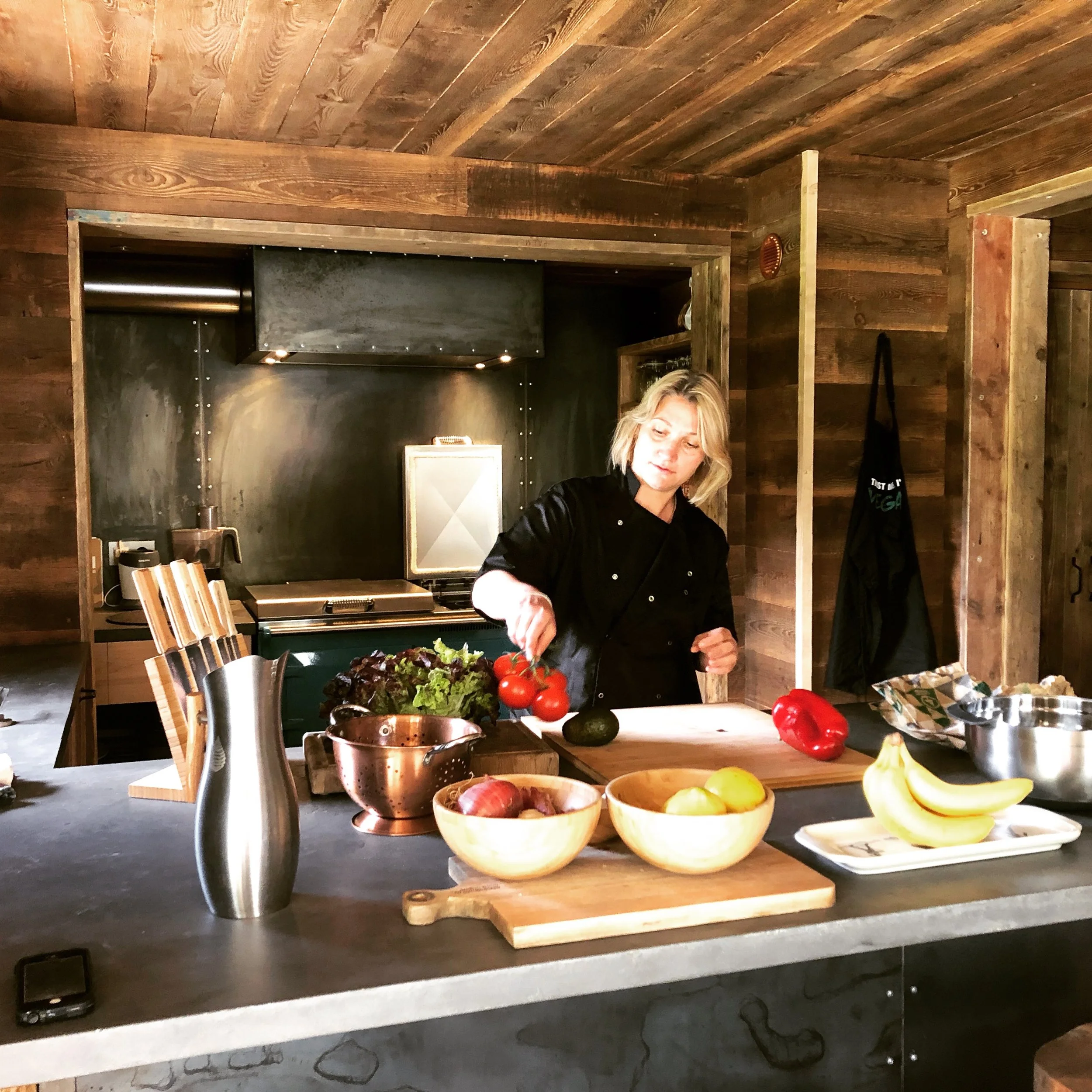 A person preparing food in a rustic wooden kitchen, with bowls of vegetables and a cutting board on a black countertop.