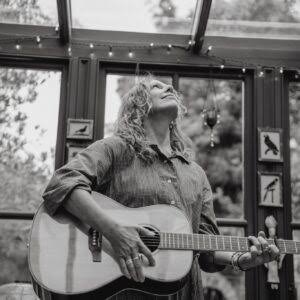Woman playing acoustic guitar during daytime indoors, windows and string lights behind her.