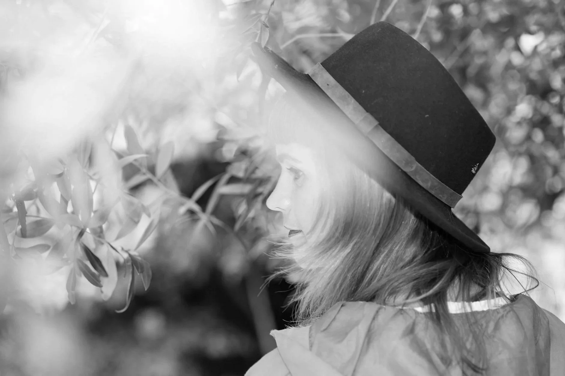 Black and white image of a doll with long hair, wearing a black top hat, looking at plants or flowers.