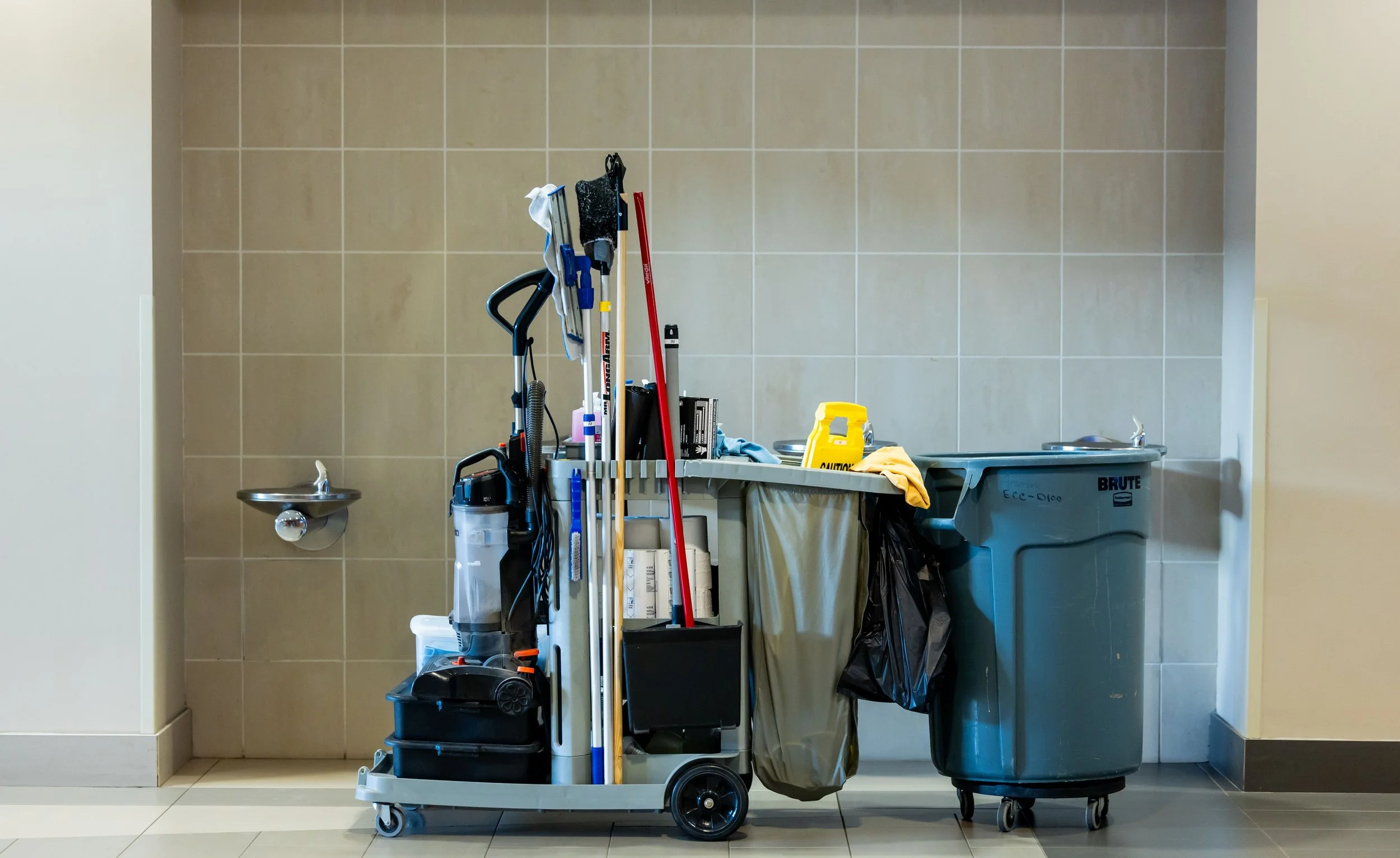 Cleaning cart filled with cleaning supplies including mops, brooms, a bucket, a spray bottle, and a trash bag next to a large trash can in a tiled hallway.