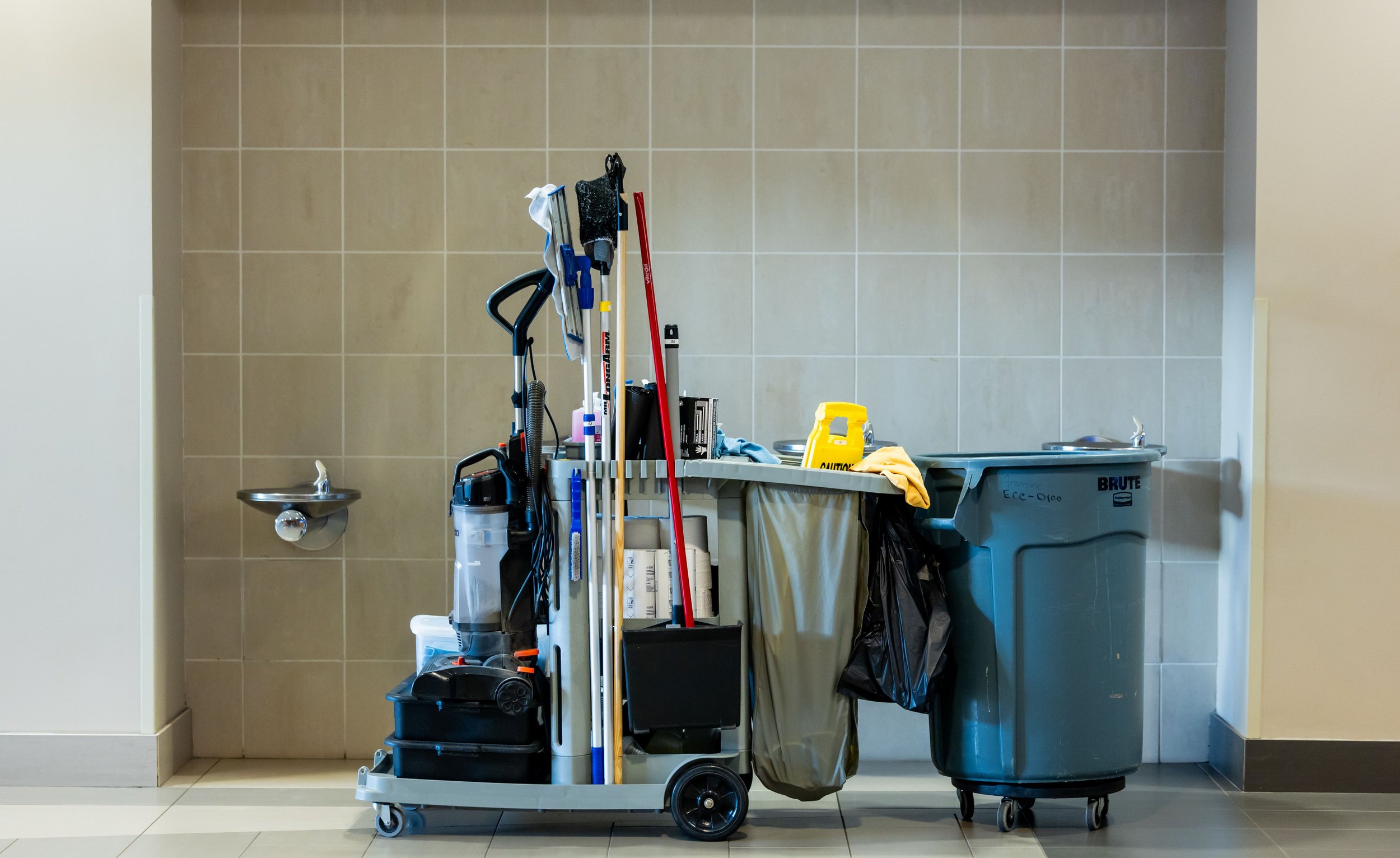 Cleaning supplies and equipment, including a mop, broom, cleaning bucket, and trash bin, arranged against a tiled wall in a cleaning area.
