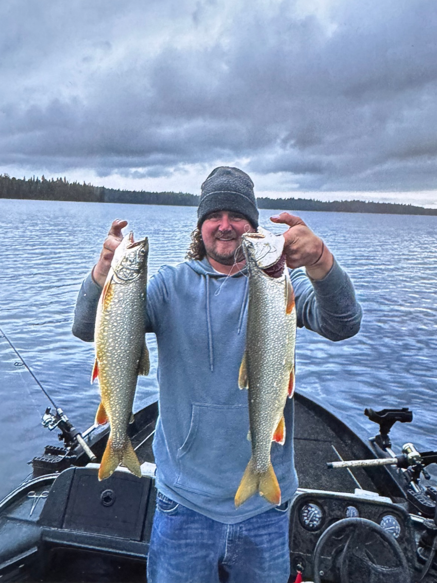 A man on a boat holding two large fish, one in each hand, with water and a cloudy sky in the background.