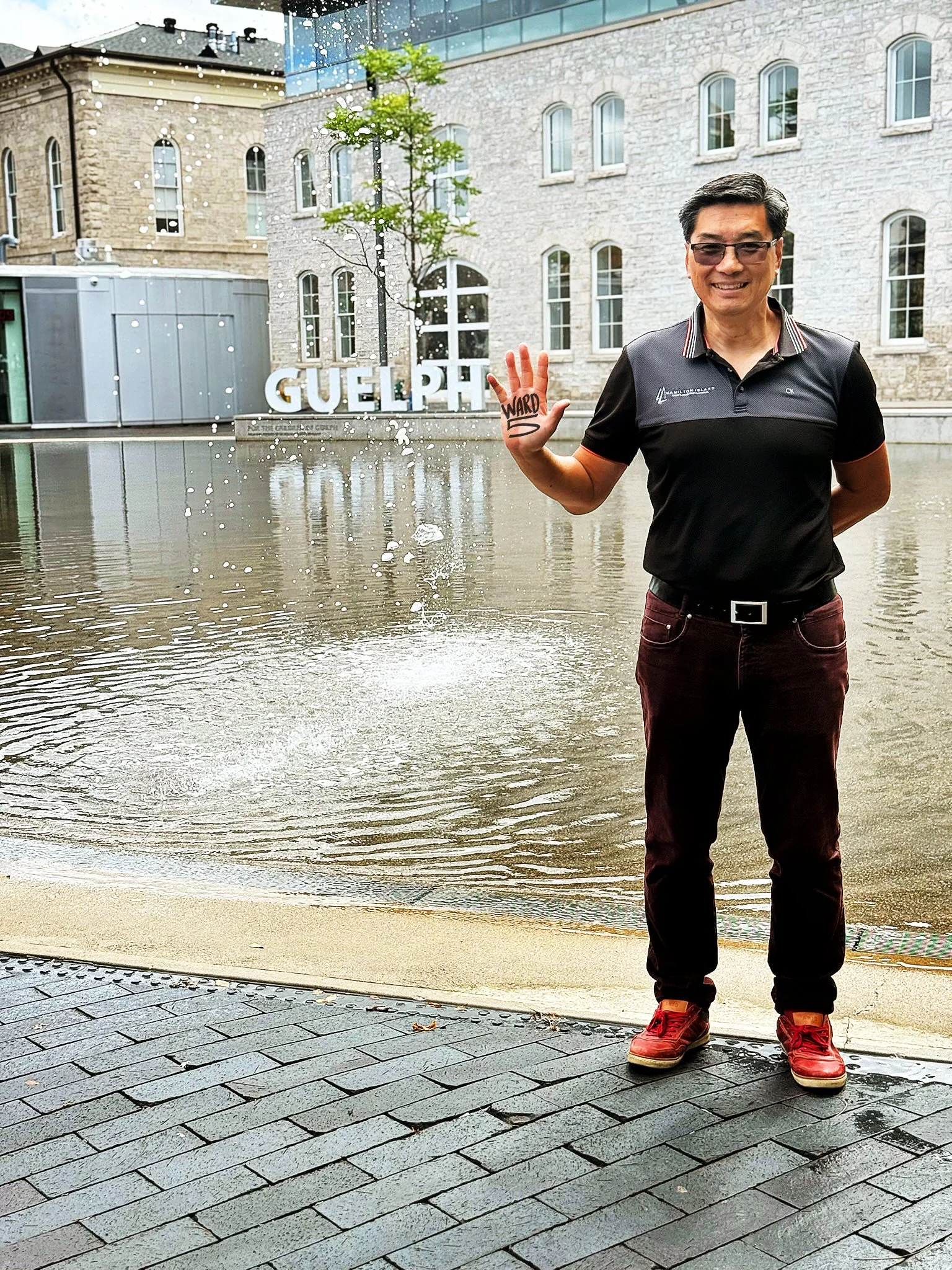 A man with glasses and black hair, smiling and waving with his right hand, stands in front of a flooded area with water covering the sidewalk. He is wearing a black polo shirt, maroon pants, and red shoes. There is a small tree and a building with arched windows in the background, and a sign that reads 'GUELP' partially visible behind the water.