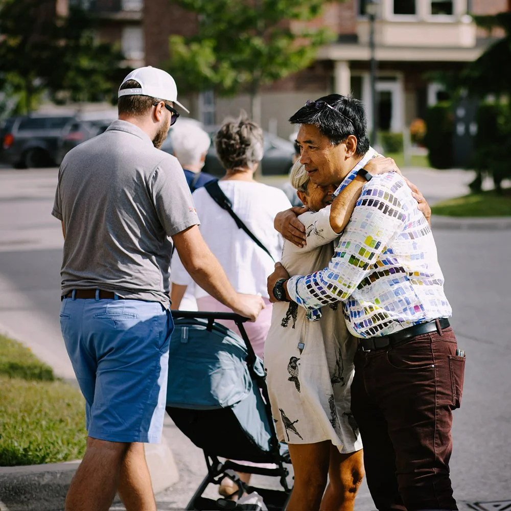 A group of people hugging outdoors on a sunny day, with a man and woman embracing in the foreground and others standing in the background near a stroller.