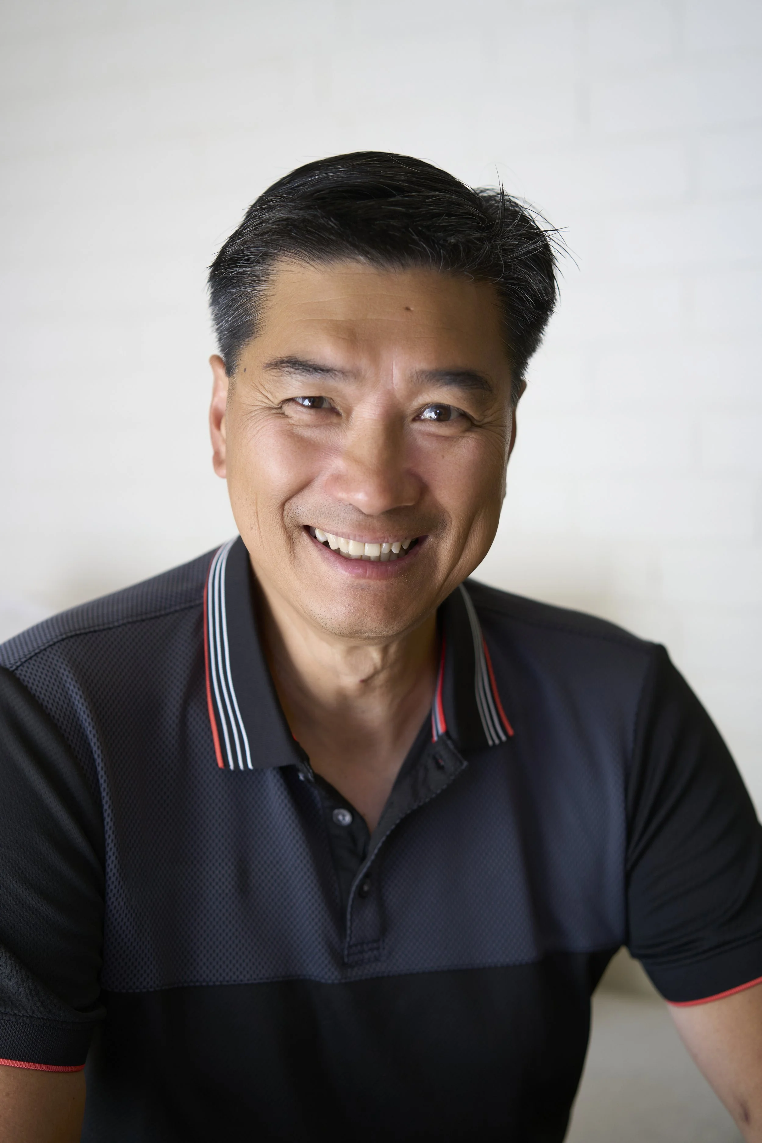 Close-up of an Asian man with dark, styled hair, smiling, wearing a black polo shirt with red and white striped accents on the collar and sleeve, against a plain light background.