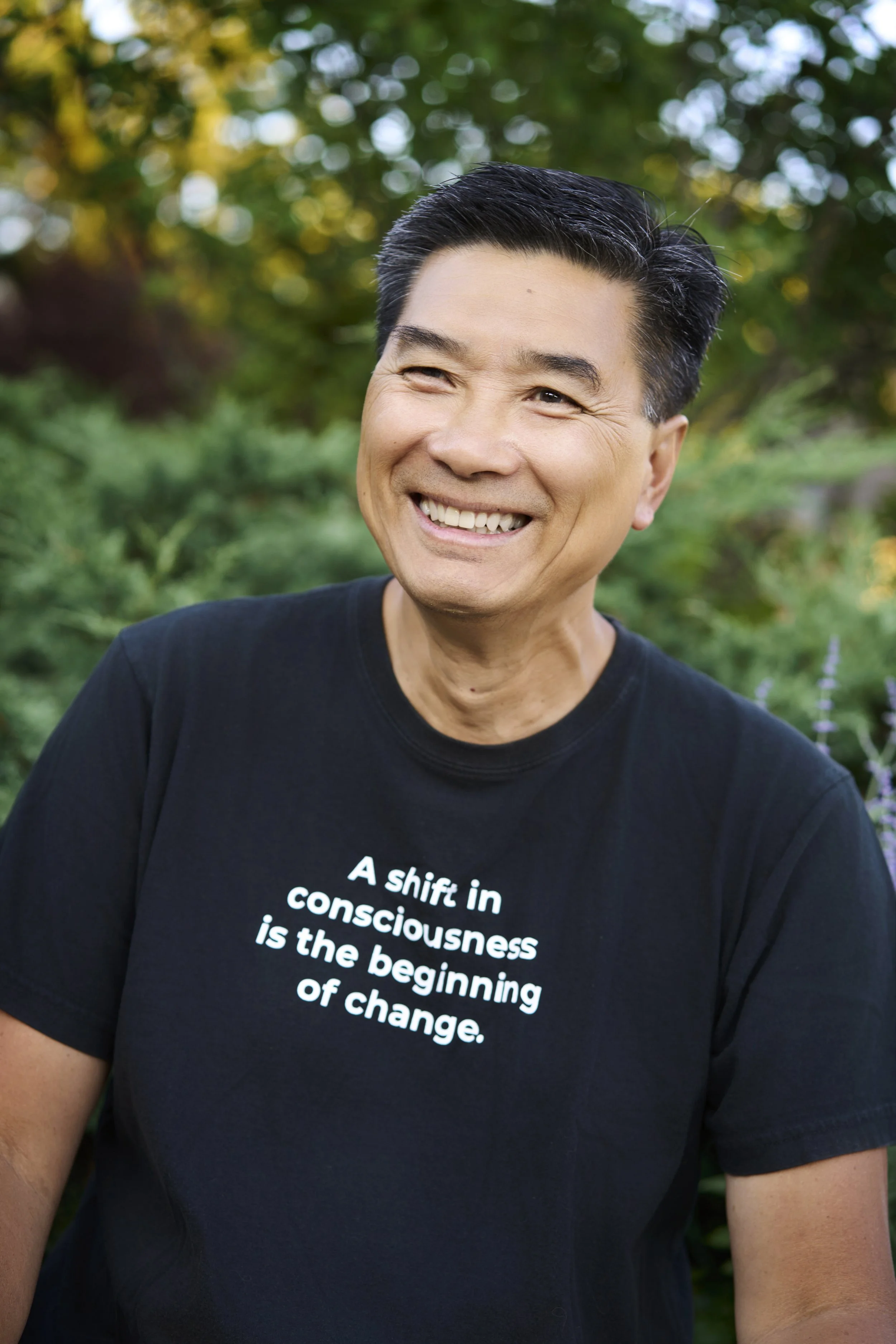 A smiling man with black hair wearing a black T-shirt with white text, standing outdoors with blurred green trees in the background.