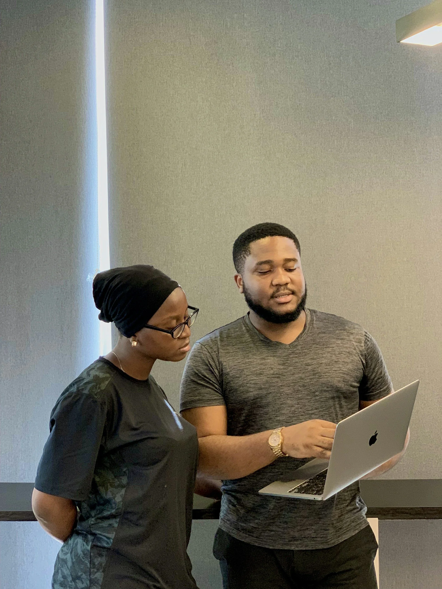 Two people standing and looking at a laptop, one in a black shirt and beanie, the other in a gray shirt, in a room with gray walls and a ceiling light.