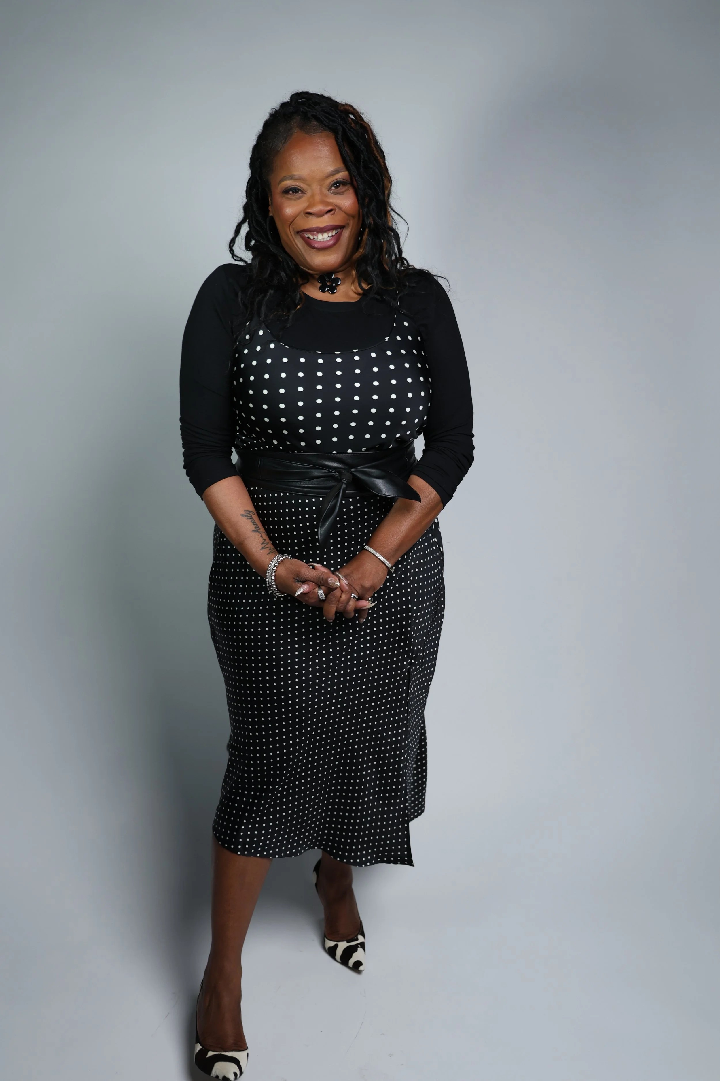 A woman with dark, curly hair styled in twists, wearing a black and white polka-dotted dress with a black belt, standing against a plain, light-colored background, smiling with her hands clasped in front of her.