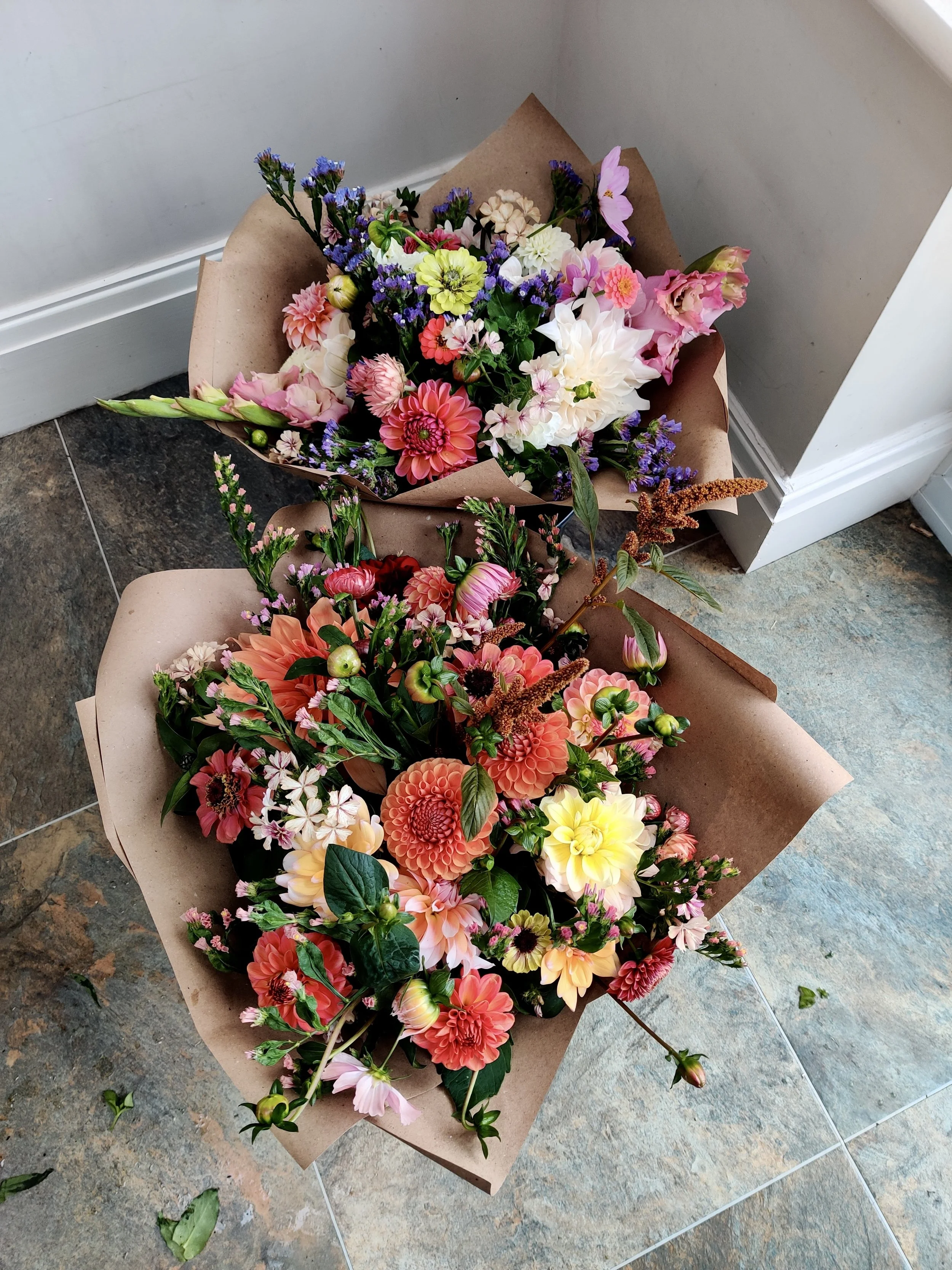Colorful bouquet of flowers including dahlias, lilies, and other mixed blooms wrapped in brown paper placed on a tiled floor near a wall.