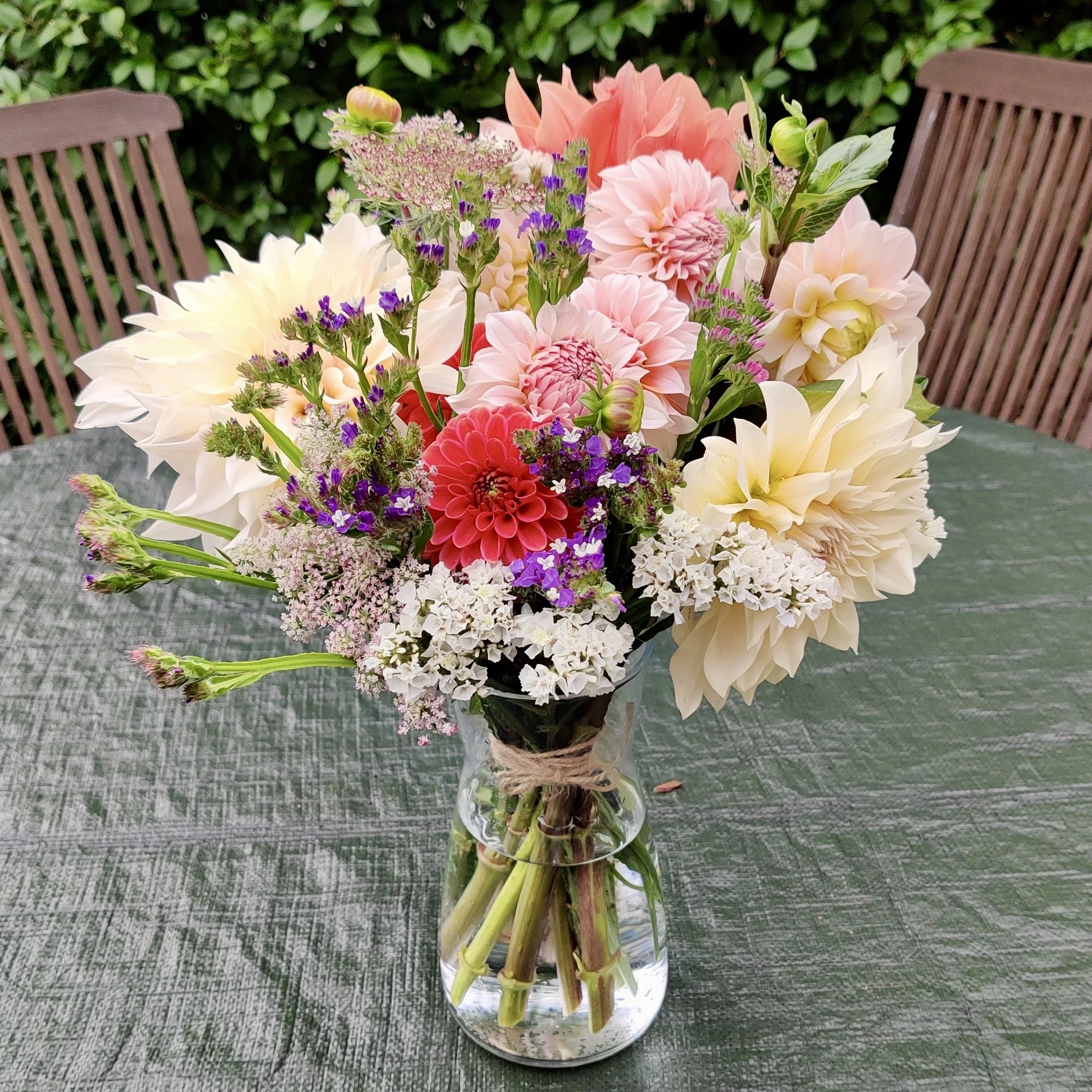 A bouquet of various colorful flowers in a glass vase on a table with a garden in the background.