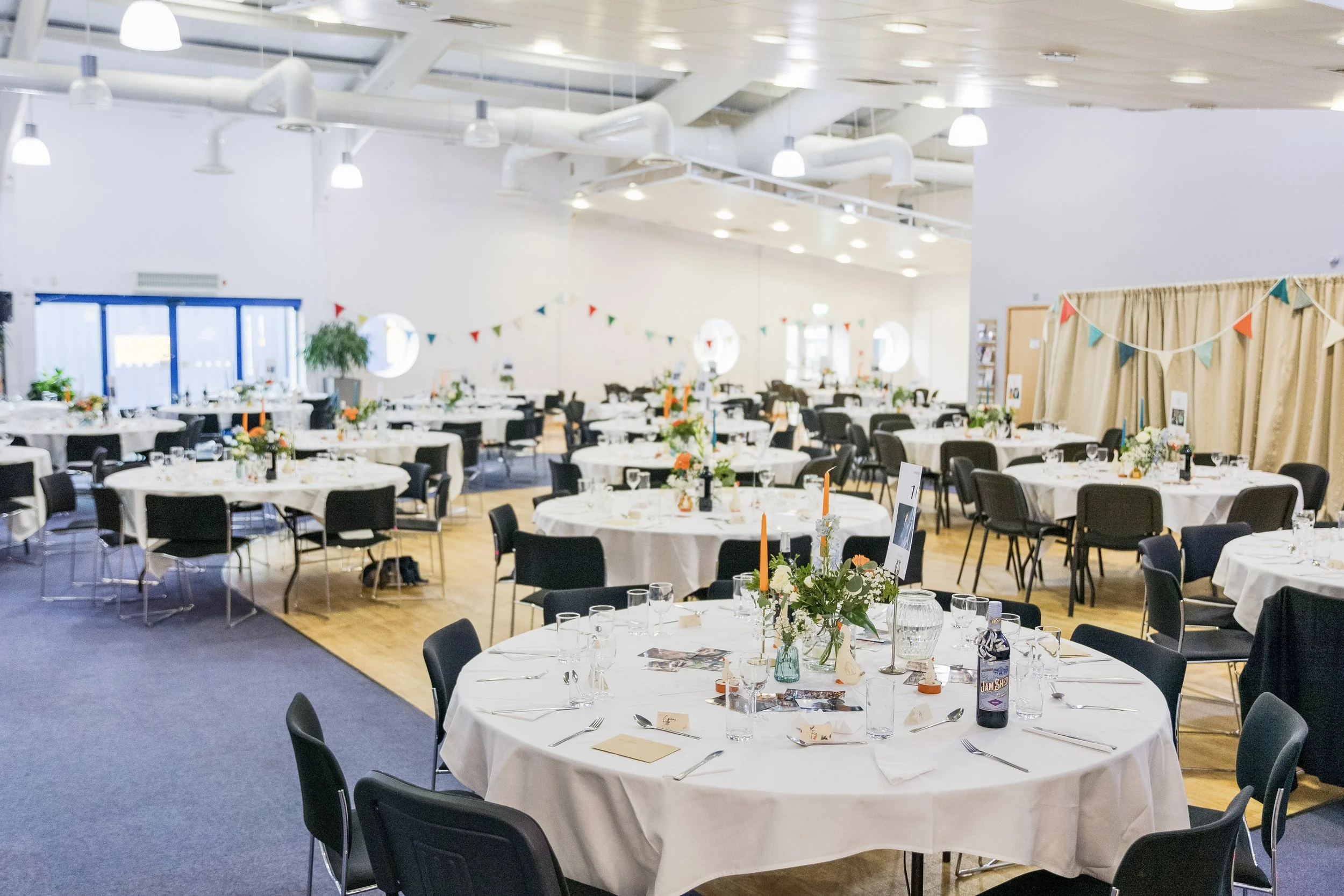 A large event space decorated with tables covered in white tablecloths, each set with glassware, plates, and silverware, and decorated with small floral centerpieces and candles. Colorful triangular pennant banners hang along the walls, and the room 