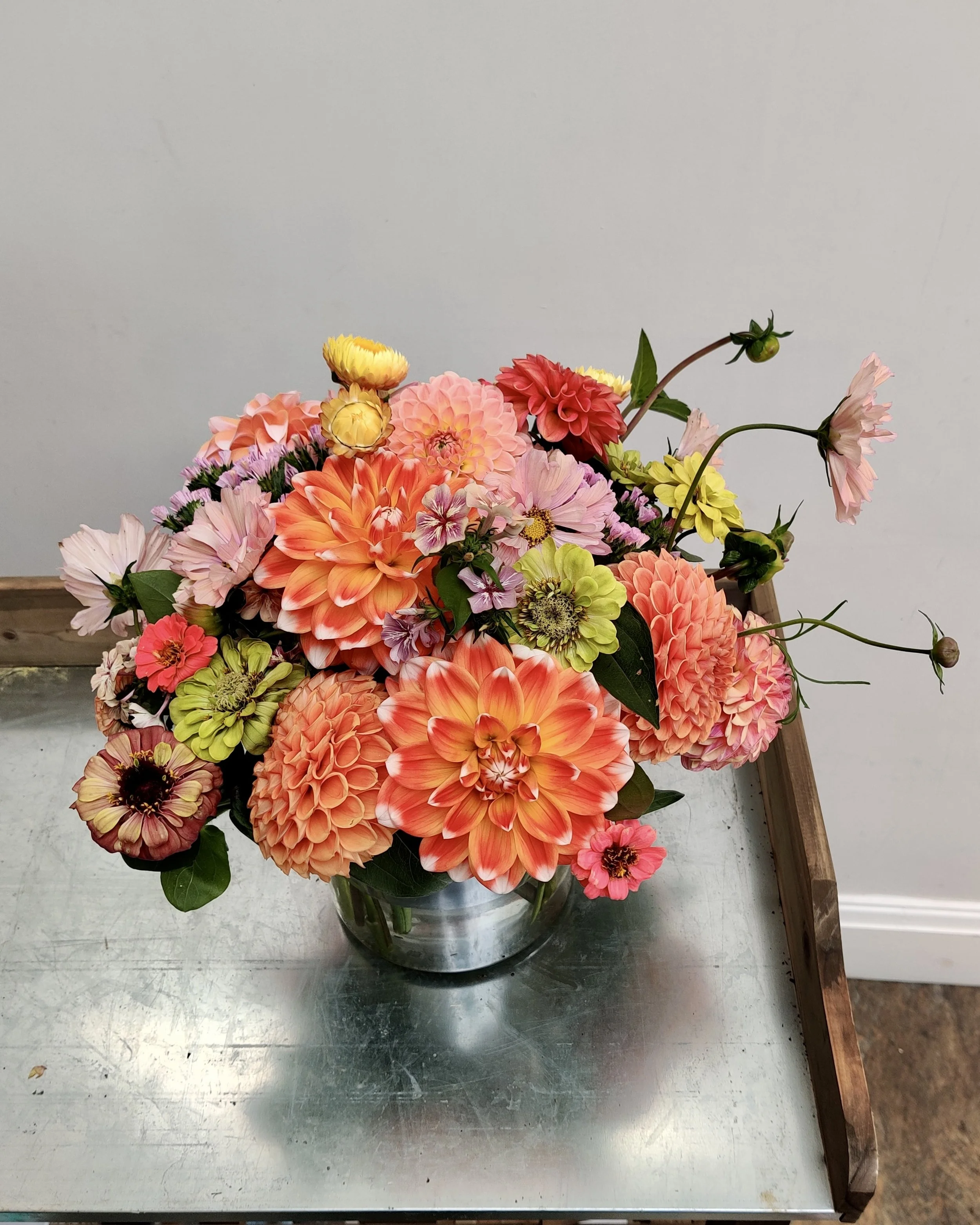 An orange, lime green and pink bouquet of various flowers, including dahlias, zinnias, and cosmos, in a glass vase on a metallic surface with a white background.