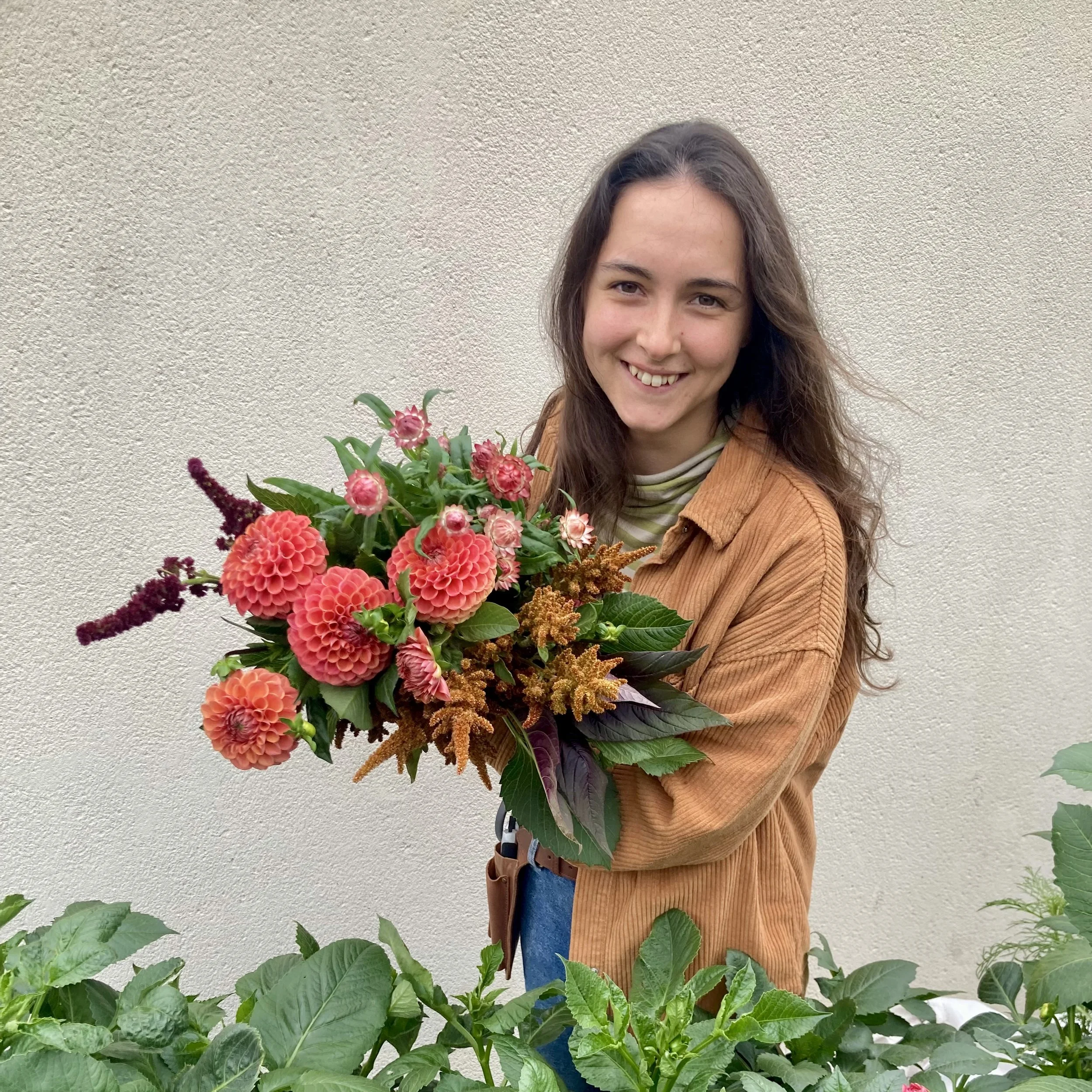 A smiling woman with long brown hair holding a bouquet of pink and red flowers, standing behind green leafy plants against a plain light-colored wall.