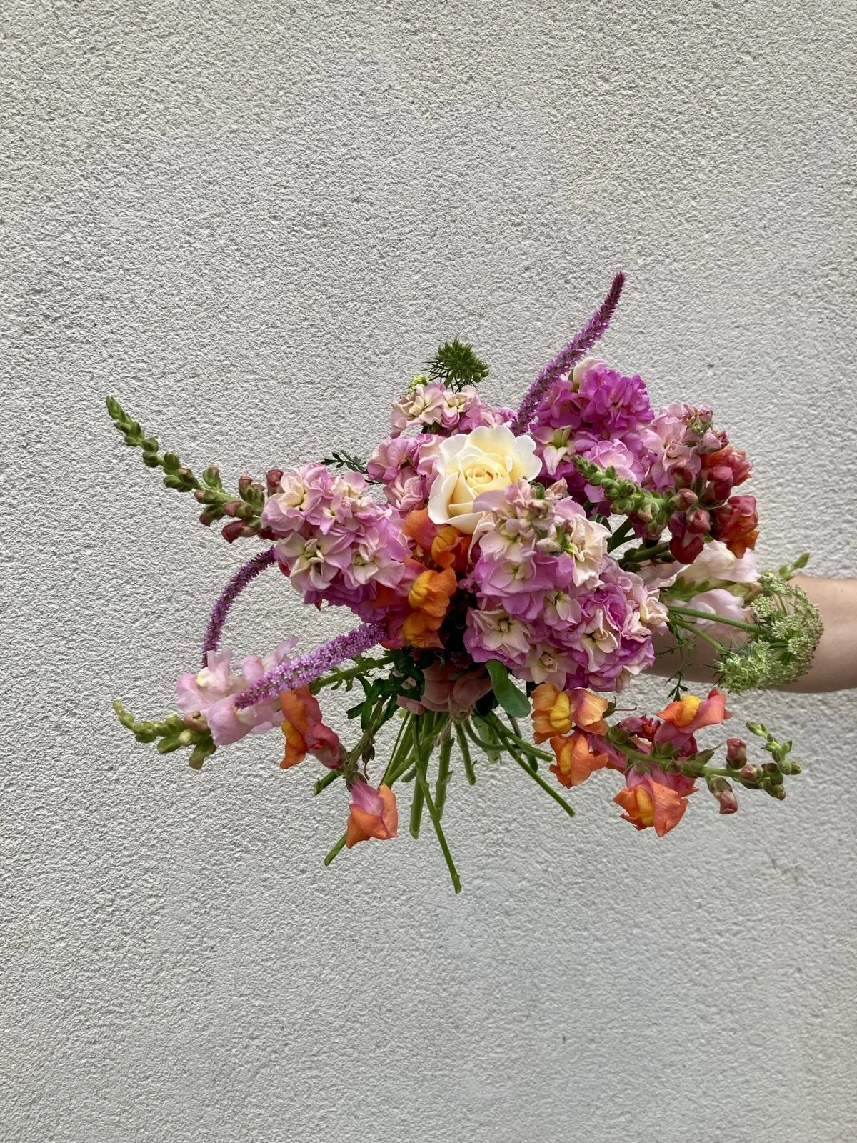 A colorful bouquet of various pink, orange, yellow, and cream flowers being held against a textured light gray wall.