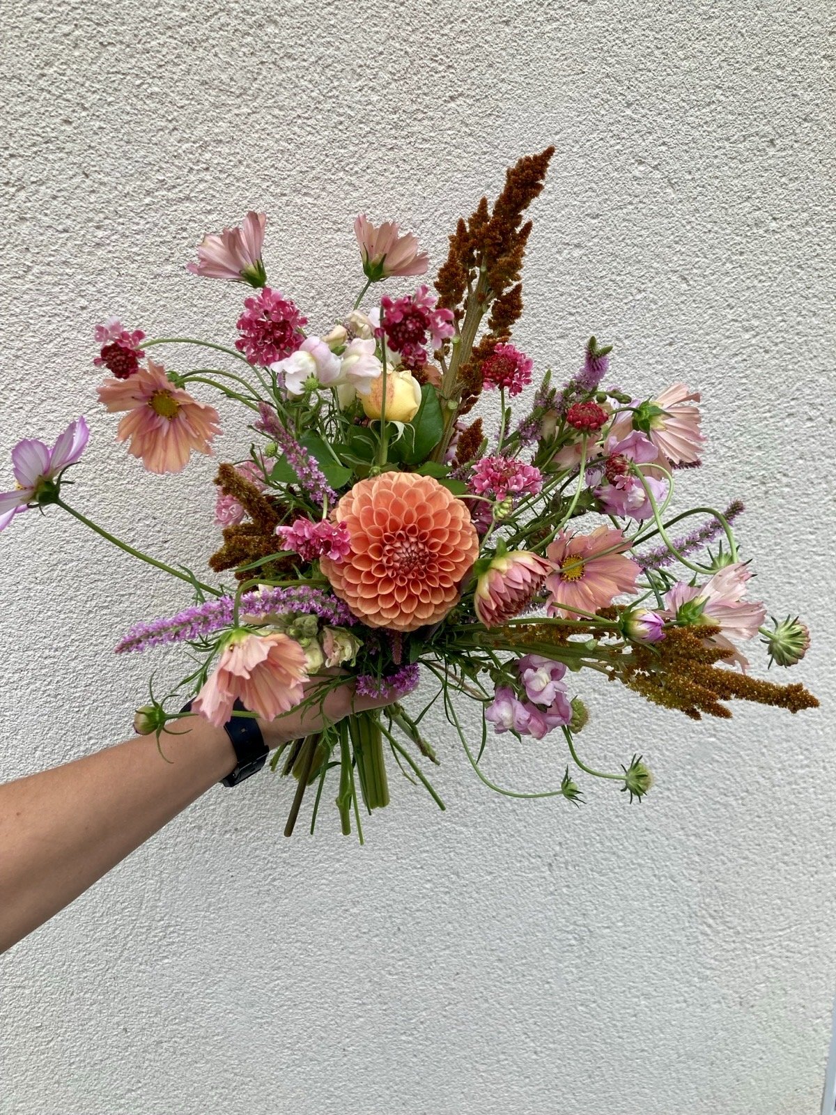 Hand holding a colorful bouquet of various pink, peach, and purple flowers against a beige textured wall.
