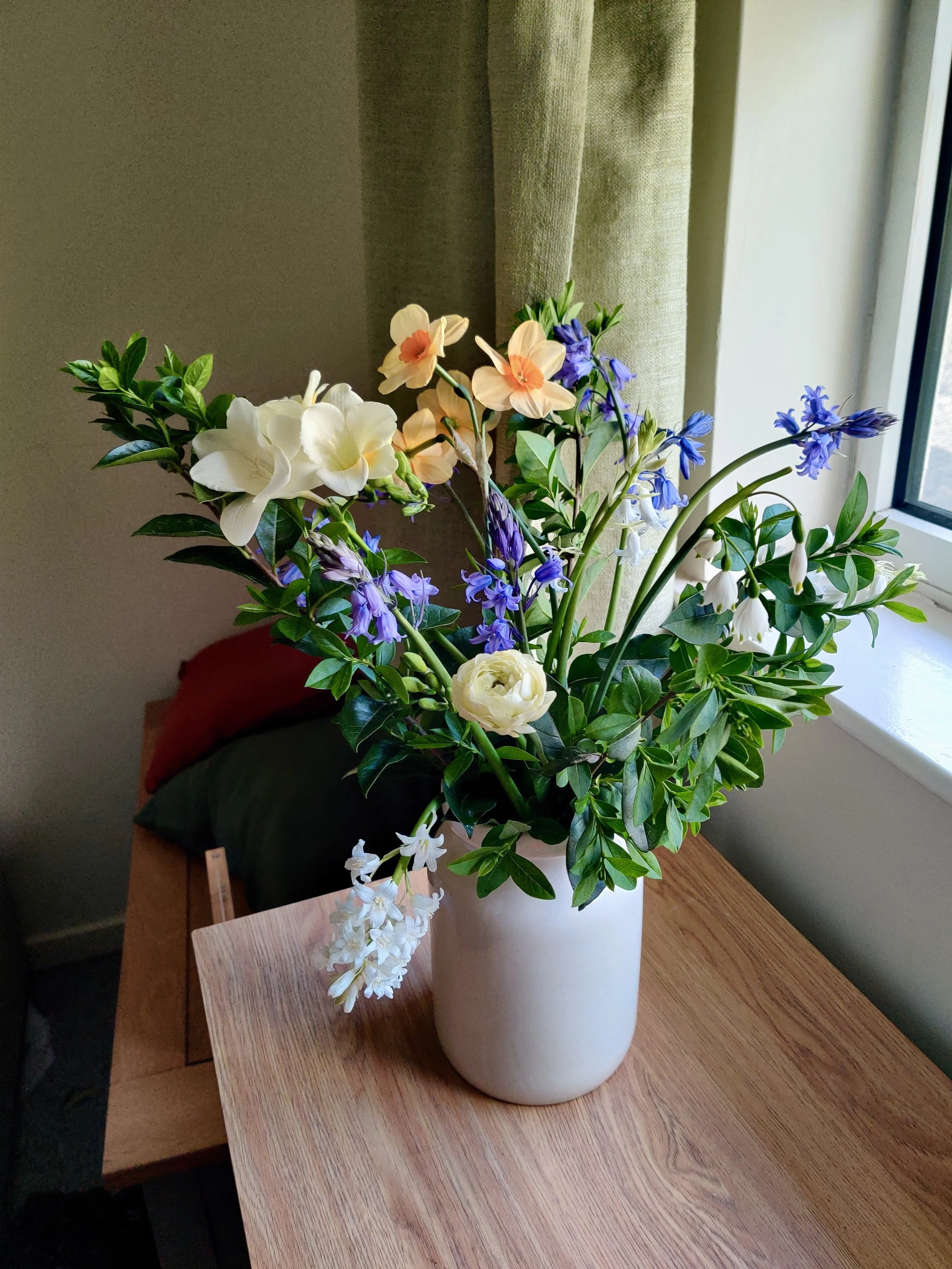 A bouquet of mixed spring flowers including peach daffodils, white ranunculus and bluebells arranged in a white vase on a wooden table by a window with olive green curtains.