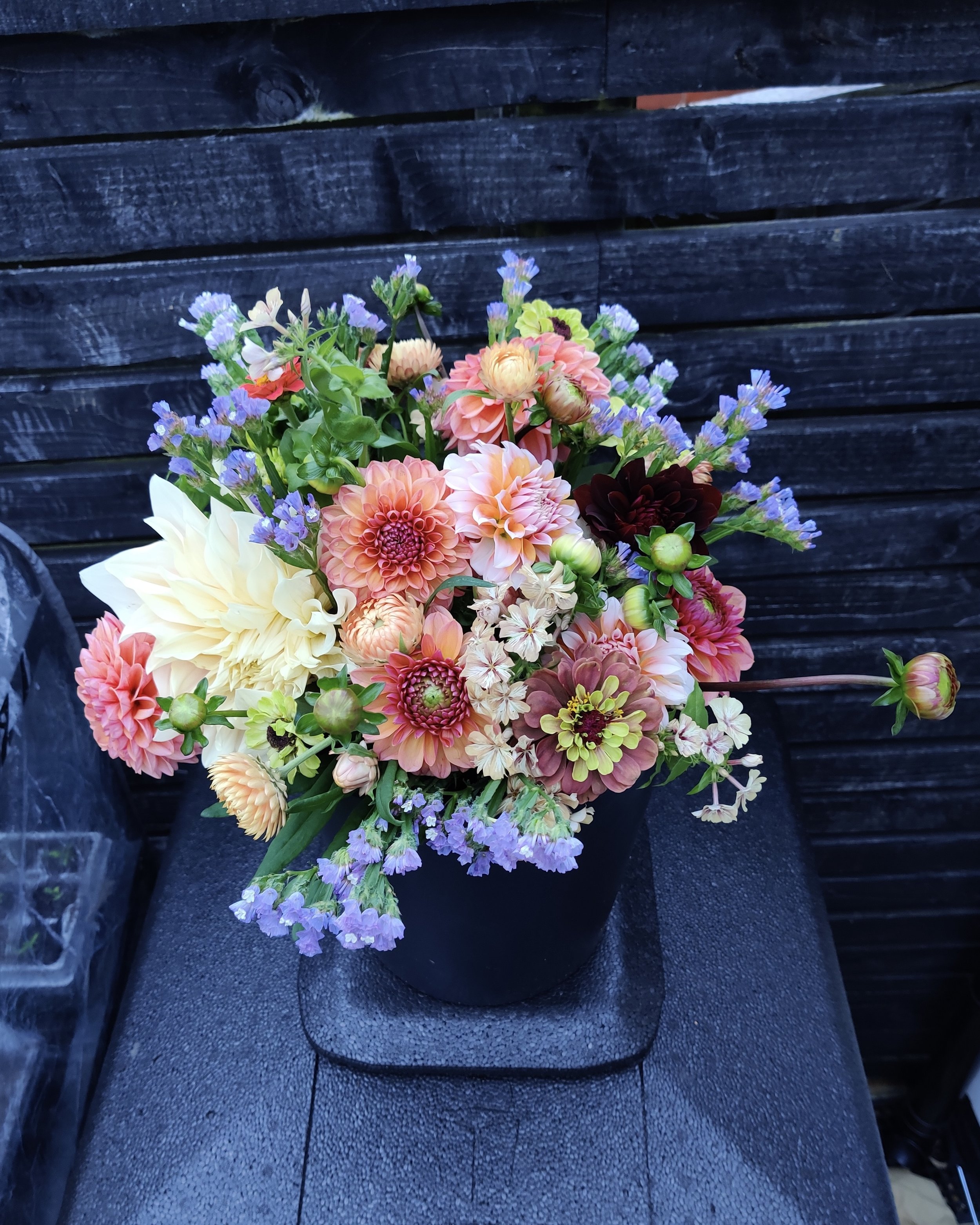 A colourful bouquet of flowers including dahlias, zinnias, statice and phlox, arranged against a dark wooden background.