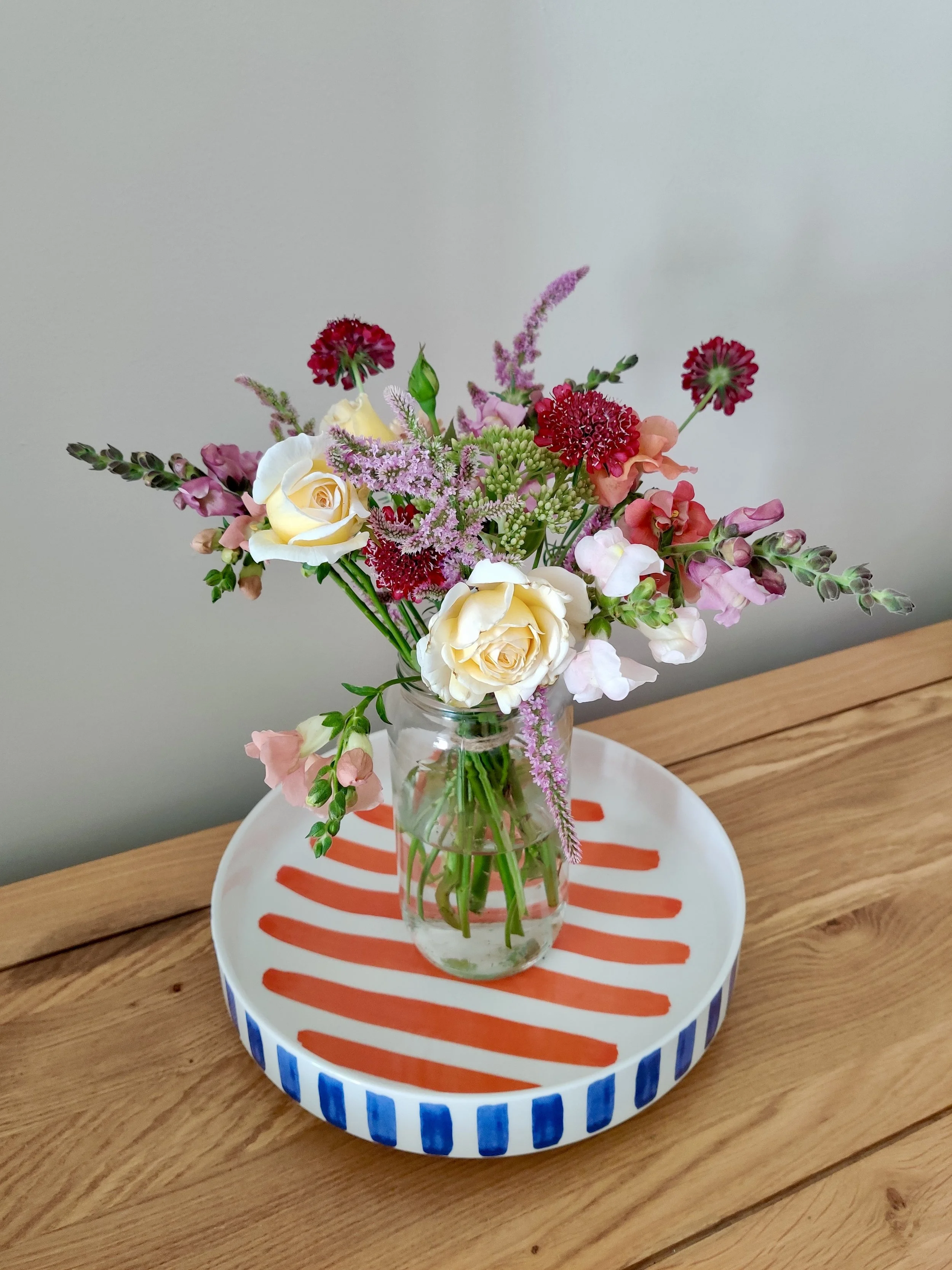 A glass vase with various colorful flowers, including white roses, pink snapdragons, purple liatris, and red flowers, sits on a round tray with red and blue stripes, placed on a wooden table against a plain light gray wall.