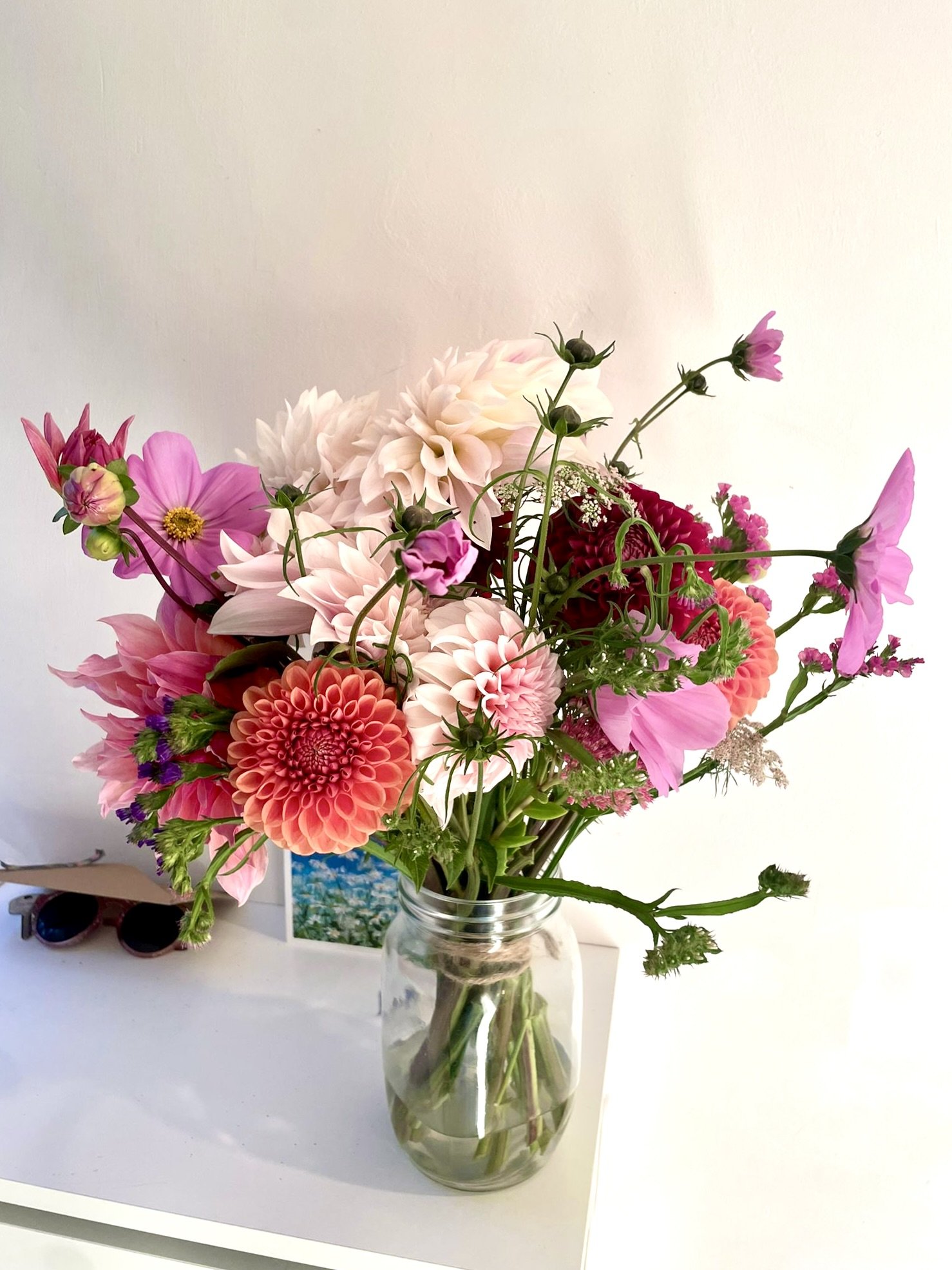 A colorful bouquet of flowers including pink, peach, white, and red blossoms in a glass vase on a white surface against a plain background.