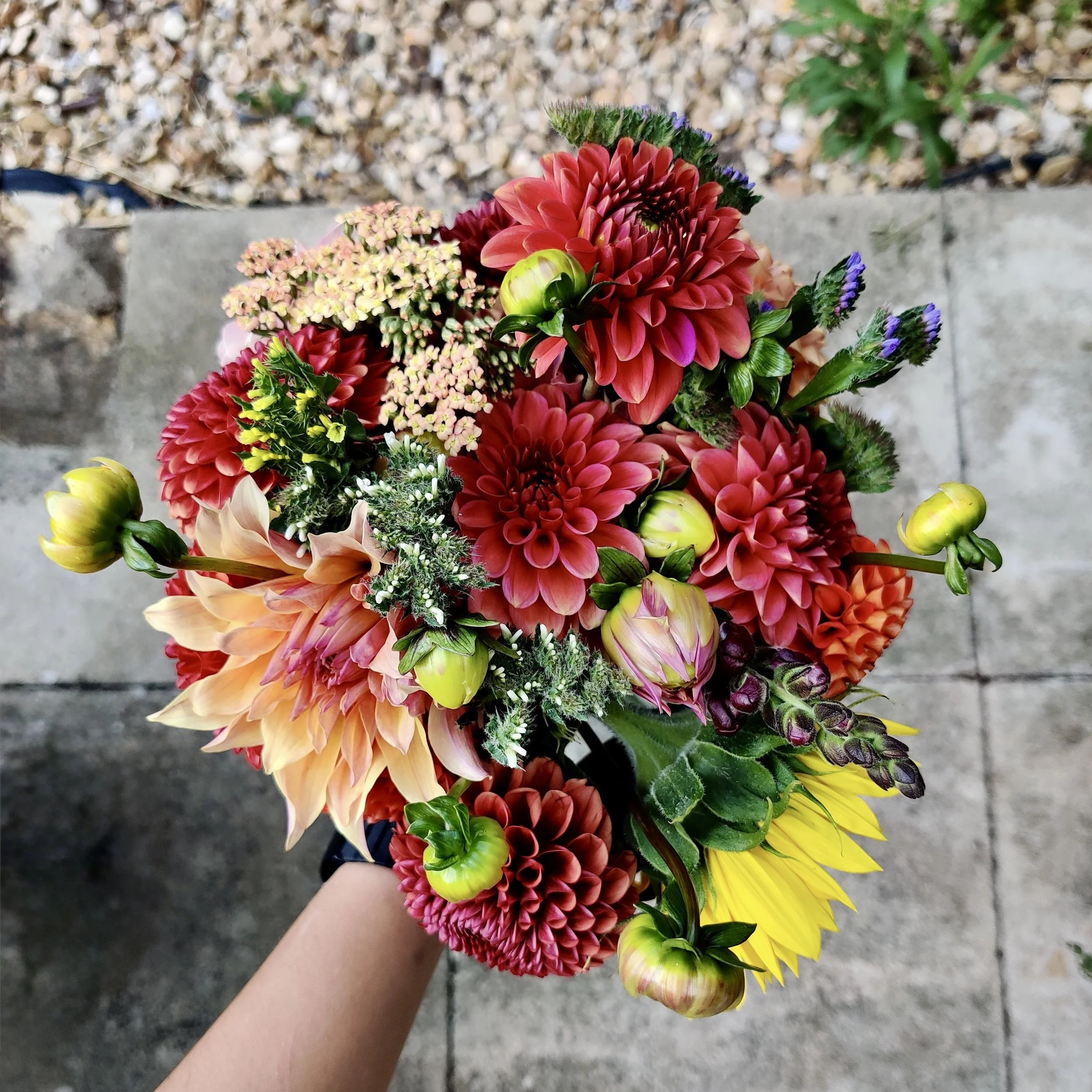 A colorful bouquet of various flowers, including red, pink, yellow, and purple blooms, held by a person's hand against a background of gravel and concrete.