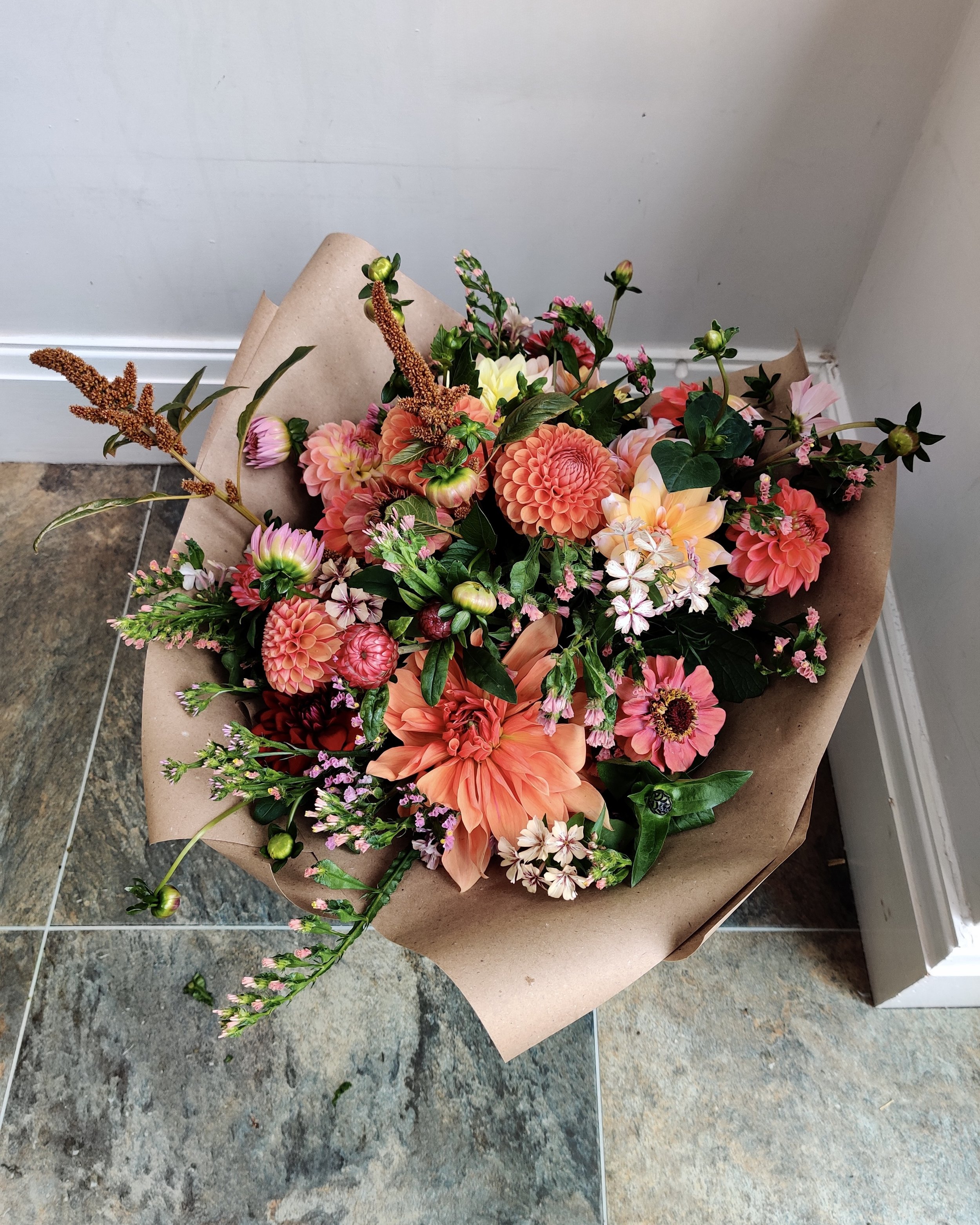 A bouquet of various colorful flowers including dahlias, zinnias, amaranth and apricot statice, wrapped in brown paper and laid on a tiled studio floor.