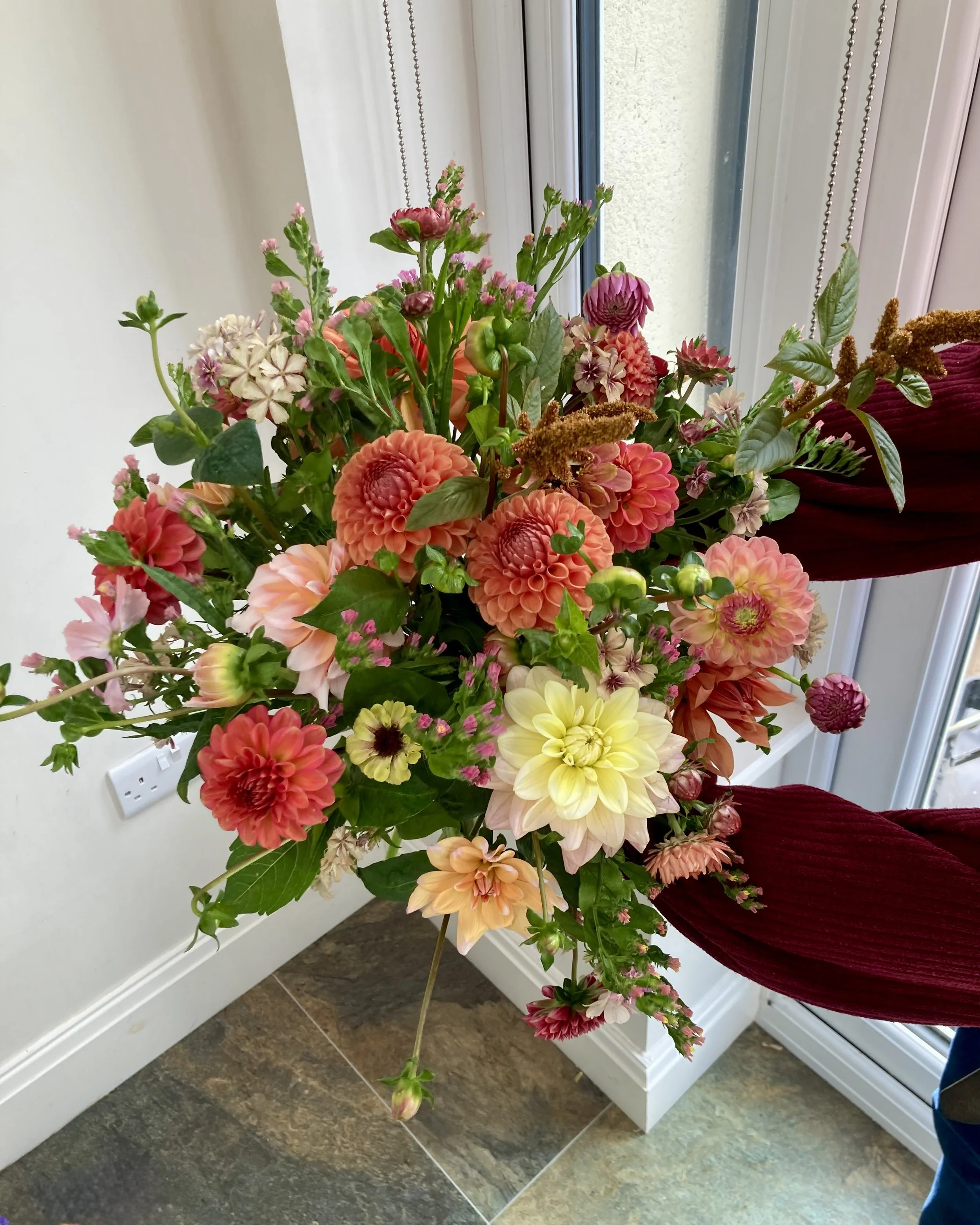 A person holding a large bouquet of mixed flowers including dahlias, zinnias, and other blooms in pink, orange, white, and yellow, standing near a white wall and window.
