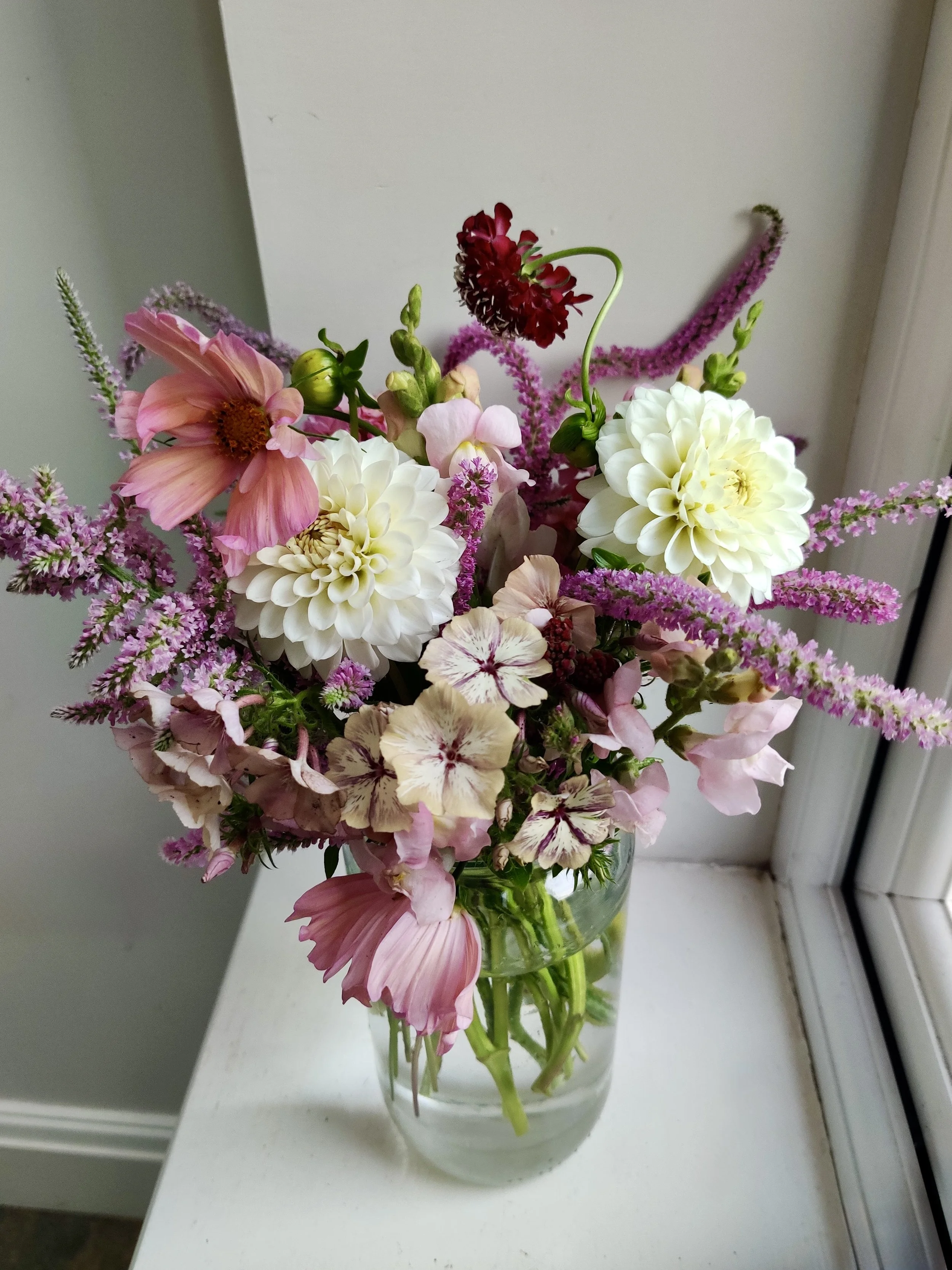 A glass vase filled with a colorful bouquet of various flowers, including pink, white, purple, and red blossoms, placed on a white windowsill.