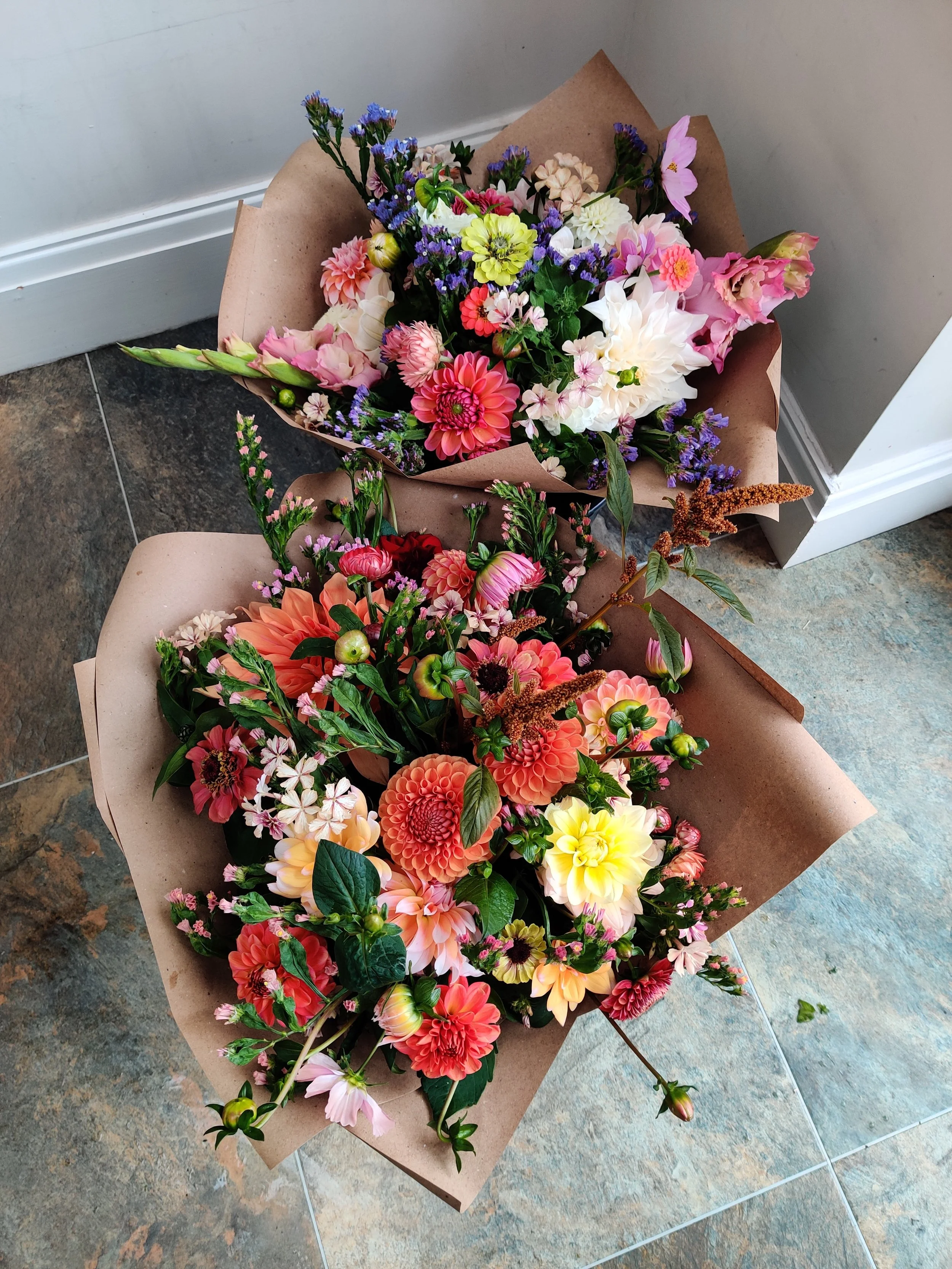 Two colourful bouquets of flowers - one peach and orange hues, one blush and blue - in brown paper floral wraps, placed on tiled studio floor.