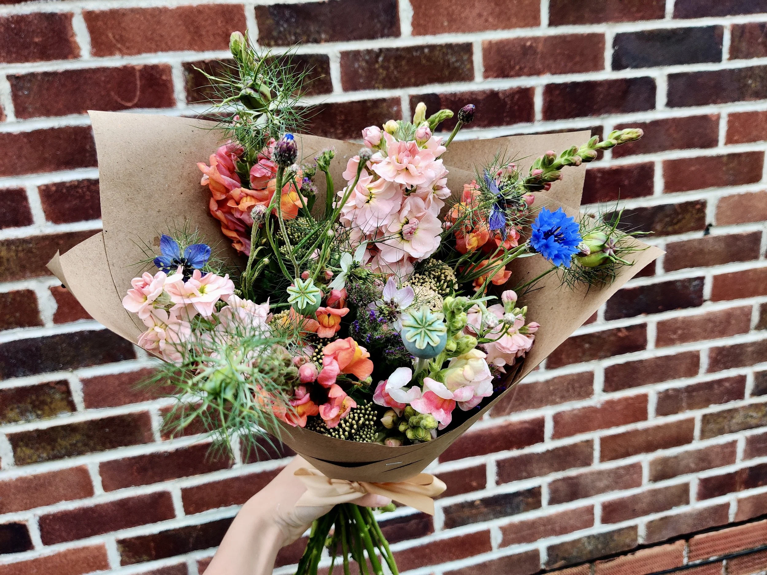 A hand holding a bouquet of colorful flowers in front of a red brick wall.
