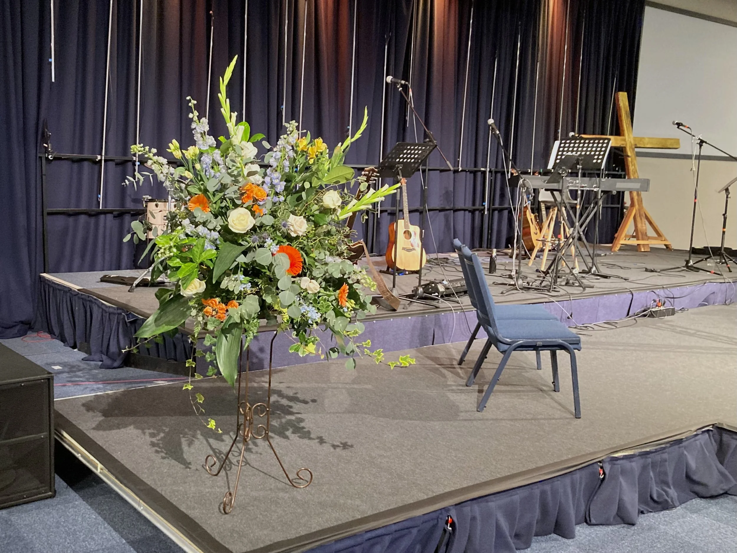 Stage with black curtains, musical instruments including guitars and a keyboard, microphones, and a large floral arrangement with white, purple, orange, and yellow flowers and green leaves.