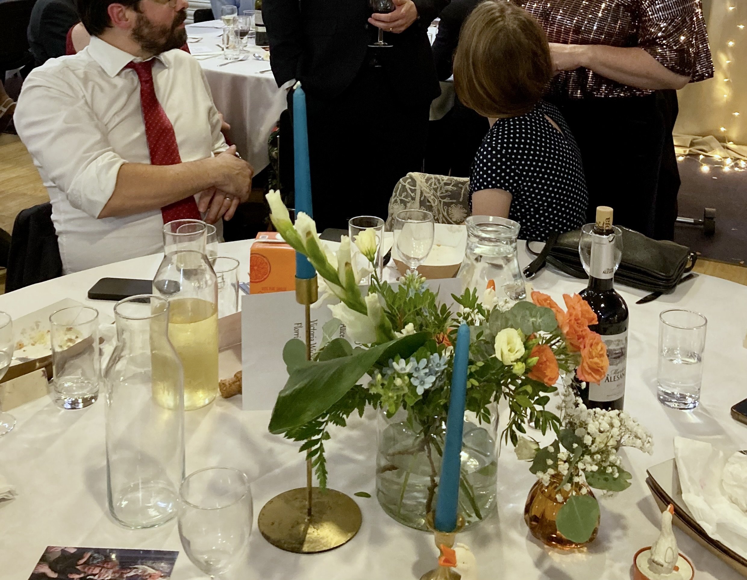 A table with flower arrangements, glasses, and bottles in a banquet hall, with people standing and sitting in the background at a social event or wedding reception.