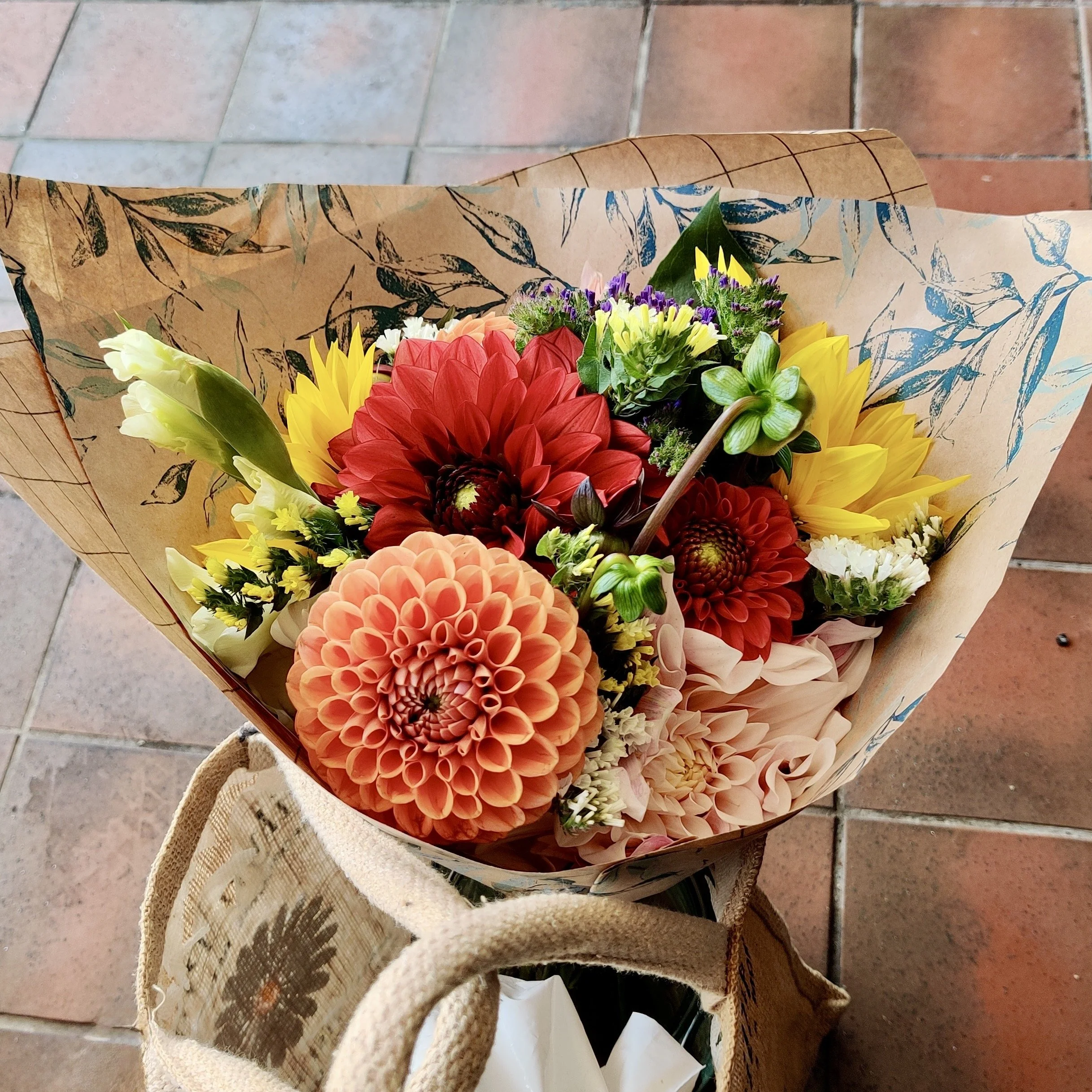 A colorful bouquet of flowers wrapped in patterned paper and a burlap cloth, placed on a tiled floor.