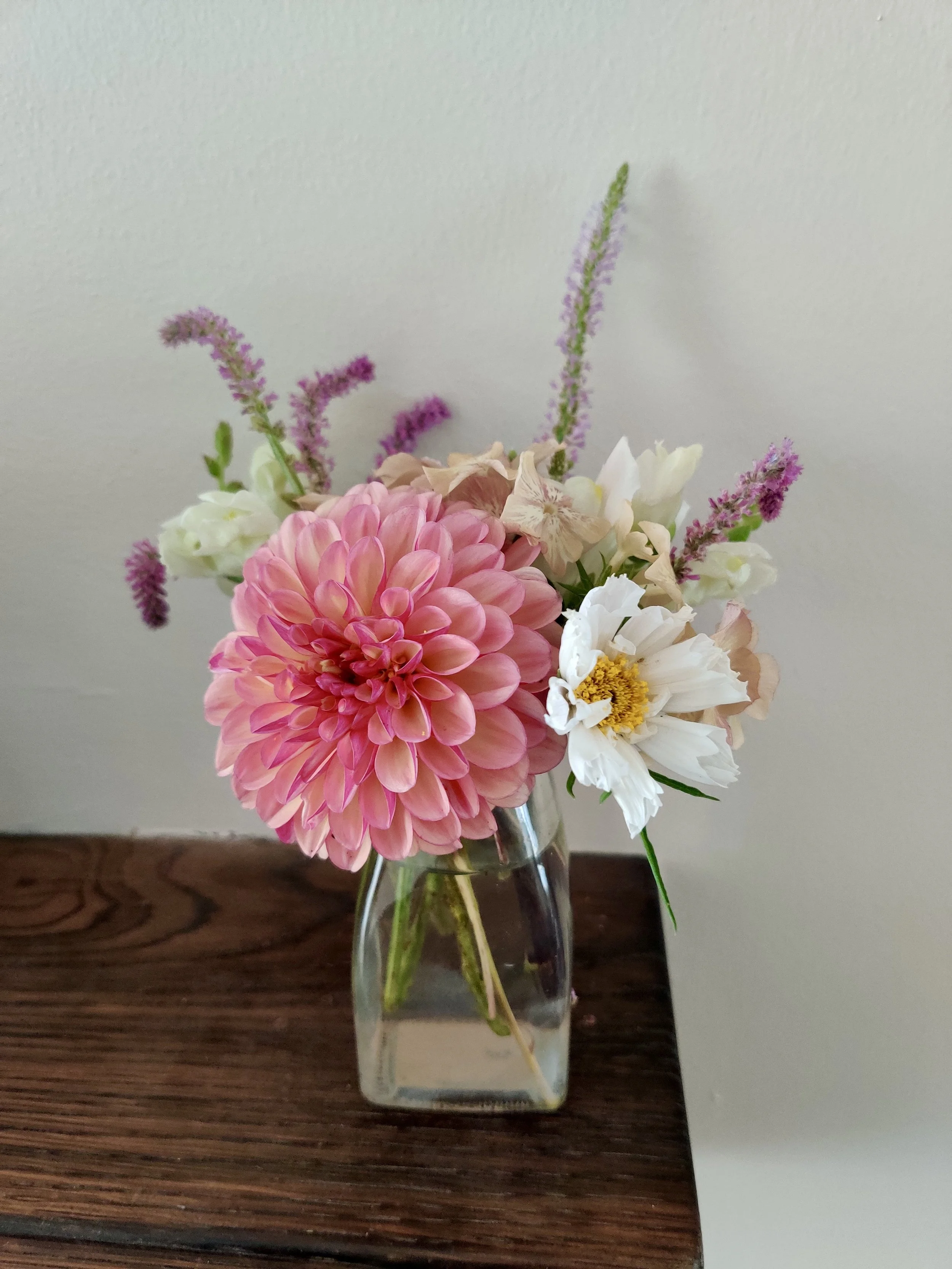 A bouquet of various flowers in a glass vase on a wooden surface against a plain wall.