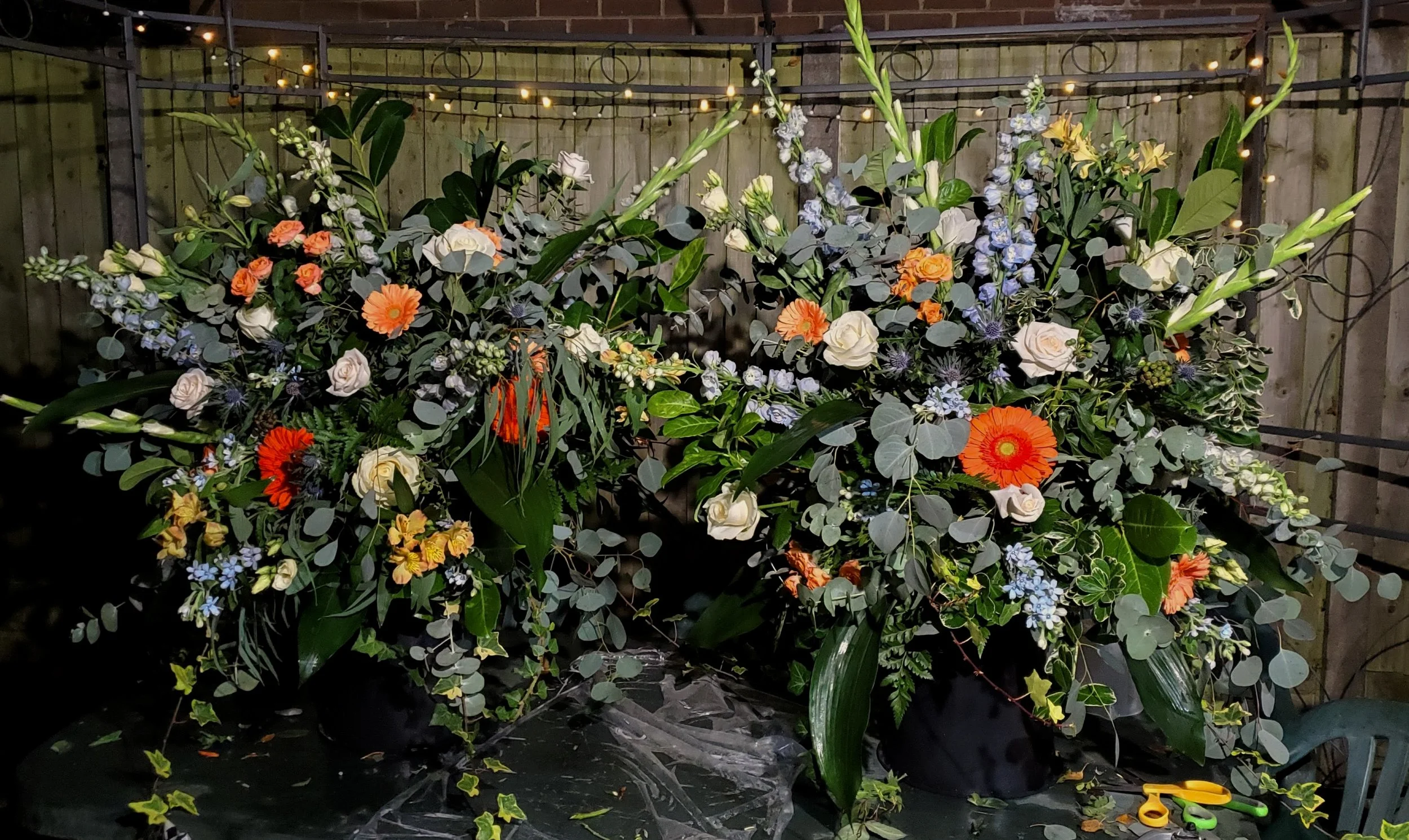 A floral arrangement with white, orange, and blue flowers, surrounded by green leaves, set against a wooden fence, decorated with string lights at night.