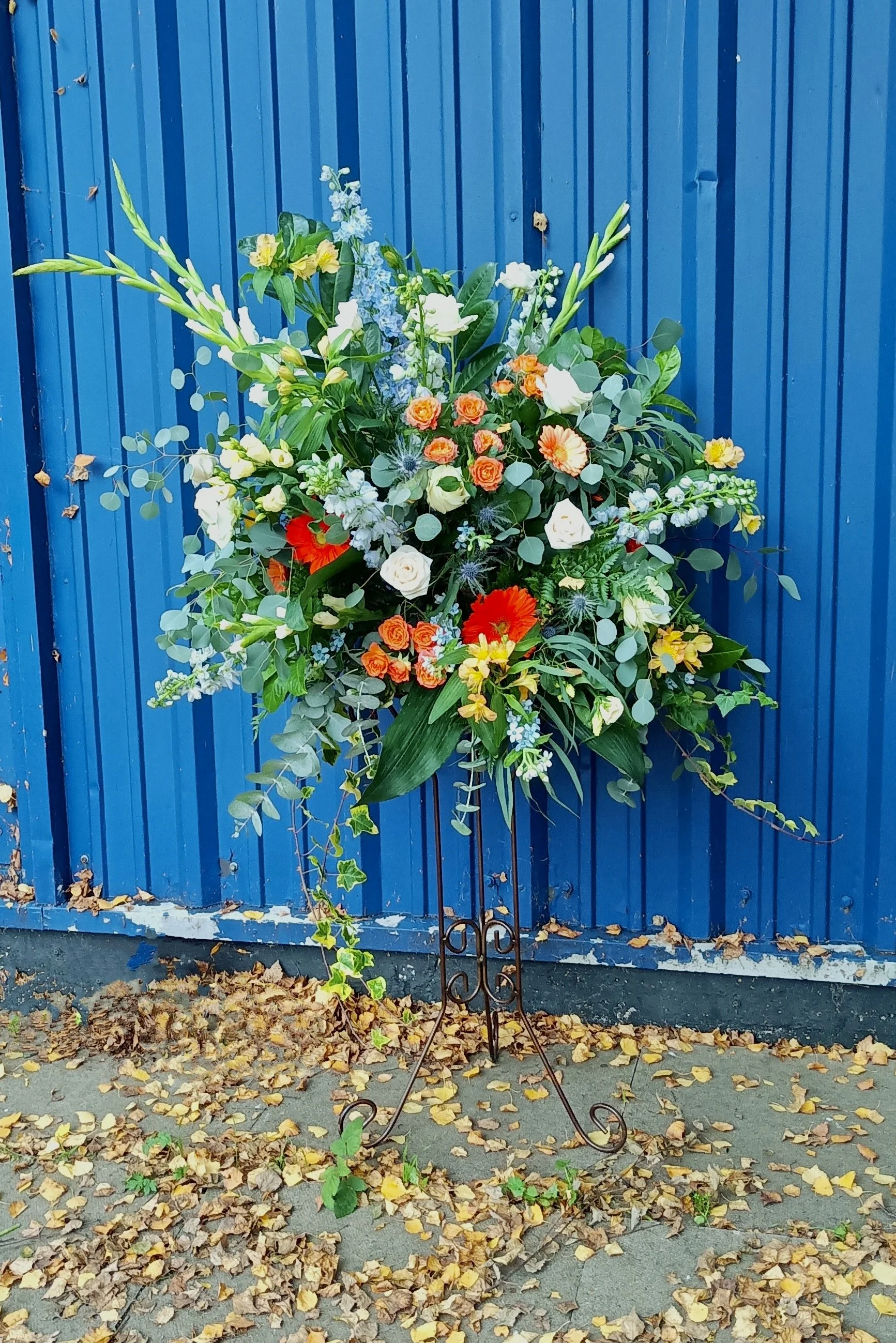 A large flower arrangement with various colorful flowers and greenery, placed in front of a blue corrugated metal fence, with fallen yellow and brown leaves scattered on the ground.