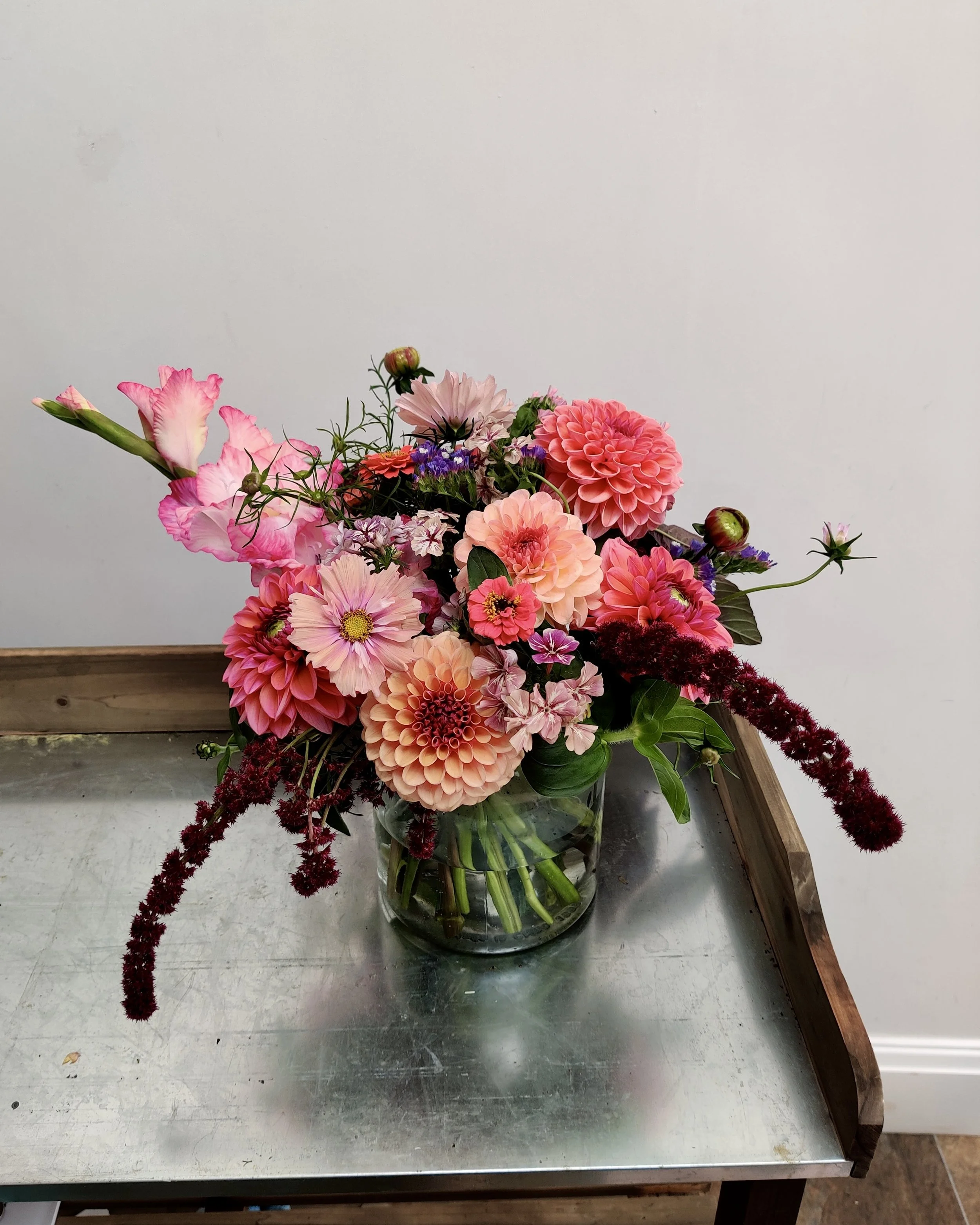 A bouquet of pink, purple, and peach flowers in a glass vase on a metal table, with a white wall and wooden ledge in the background.