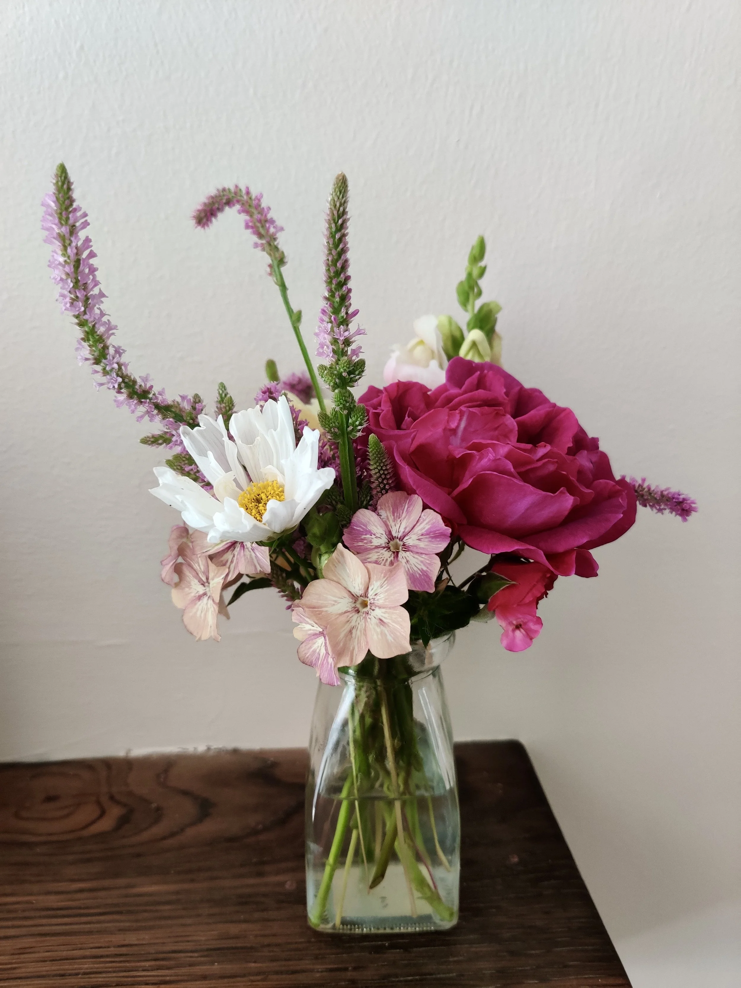 A mixed bud vase with pink, white, and purple flowers on a wooden mantlepiece against a plain wall.