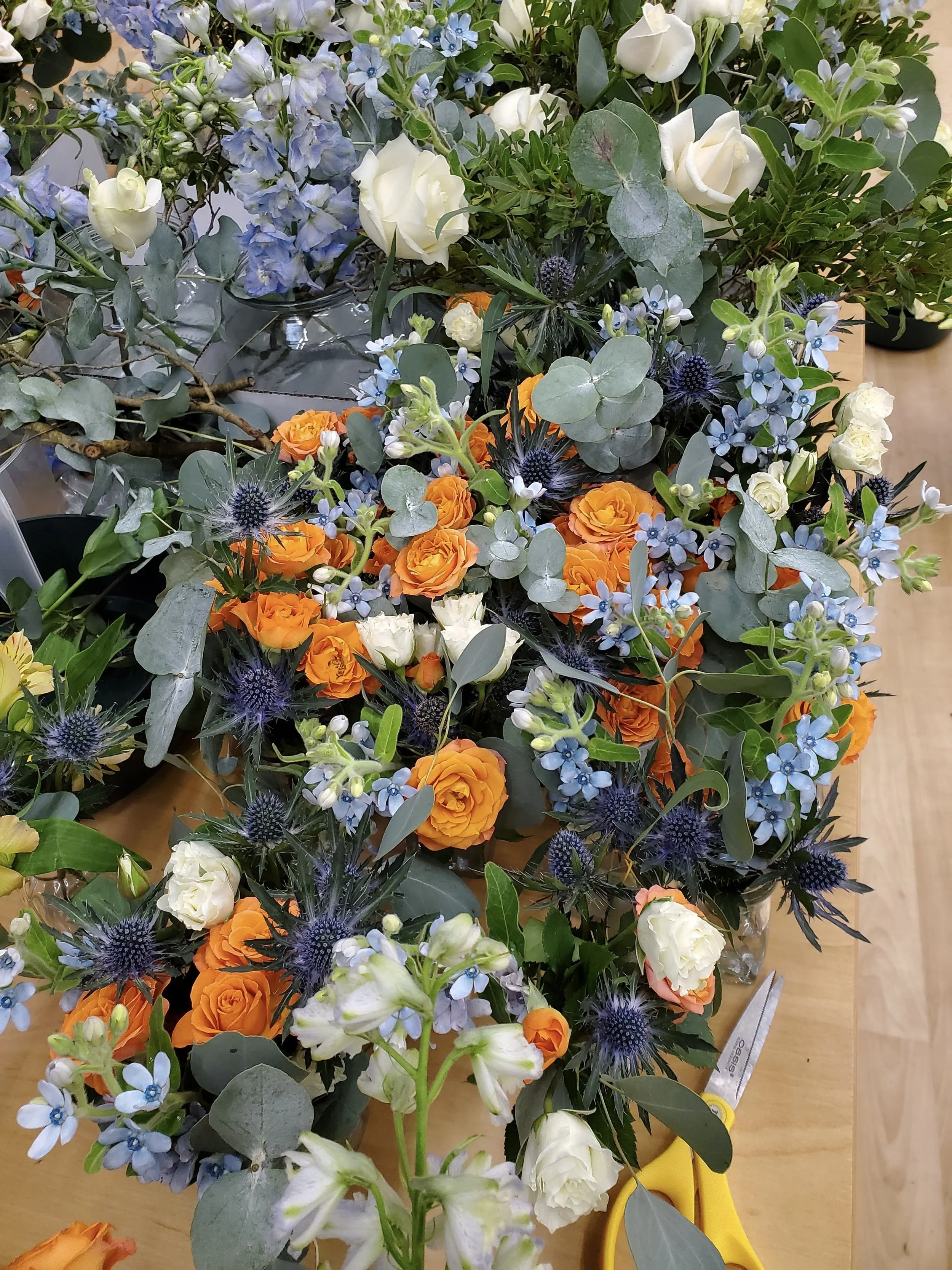 A colorful assortment of fresh flowers, including white roses, orange roses, blue delphiniums, blue thistles, and various greenery, arranged on a wooden surface with scissors nearby.
