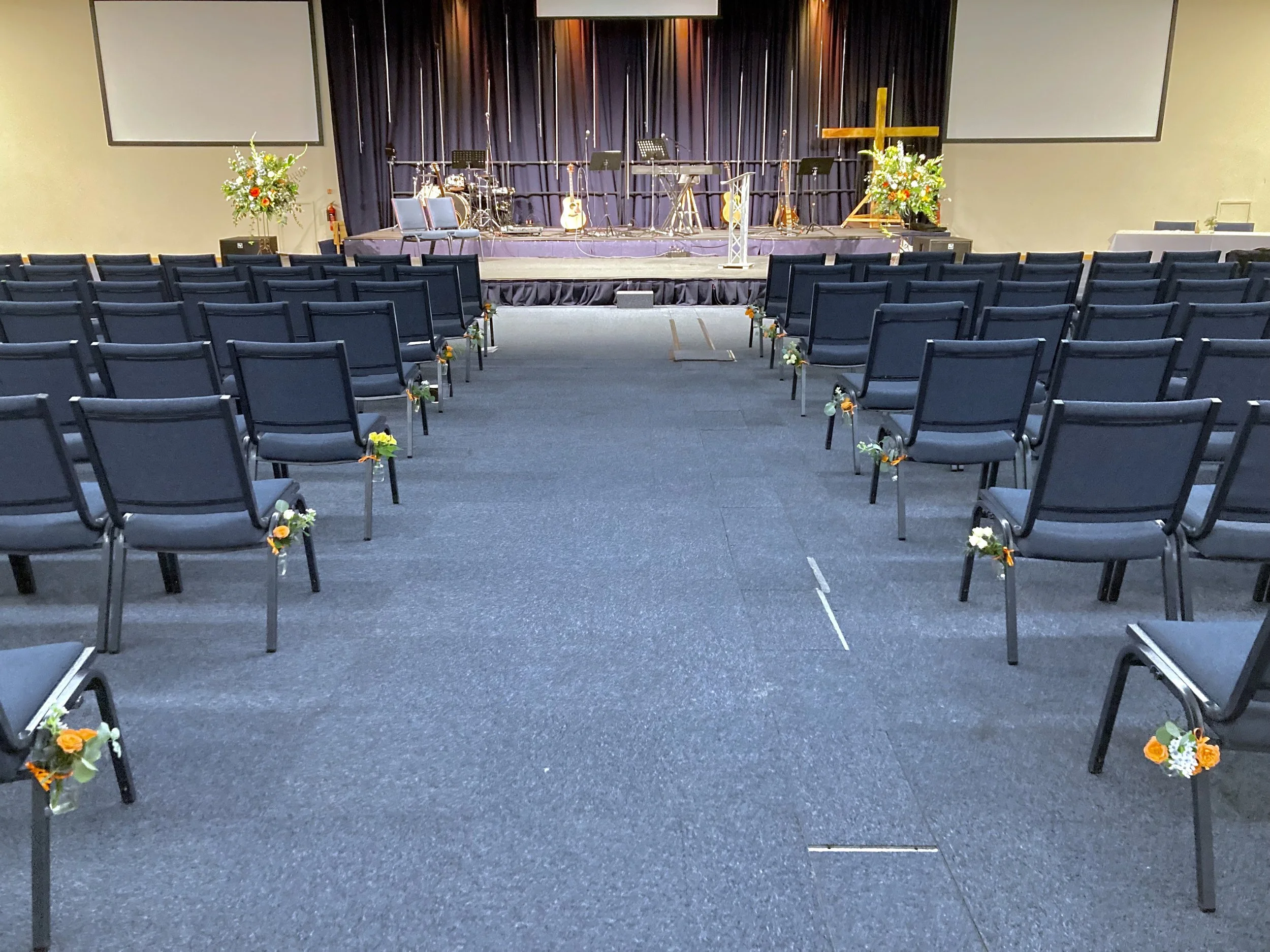 Empty church or event hall with rows of chairs decorated with small flower arrangements, a stage with musical instruments, microphones, and a wooden cross, and large screens on either side of the stage.