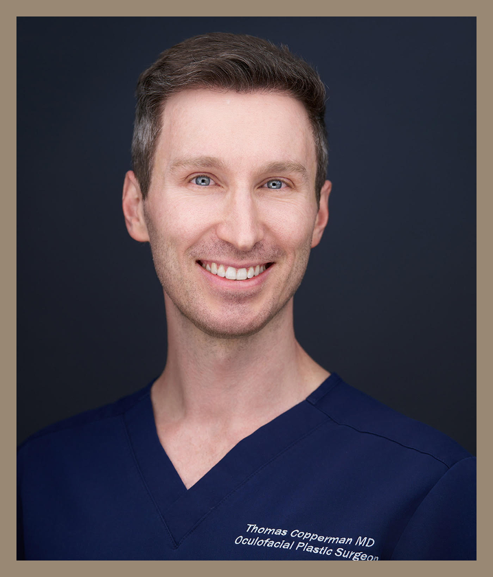 A professional headshot of Thomas Copperman MD, an oculofacial plastic surgeon, wearing blue scrubs against a dark background, smiling at the camera.