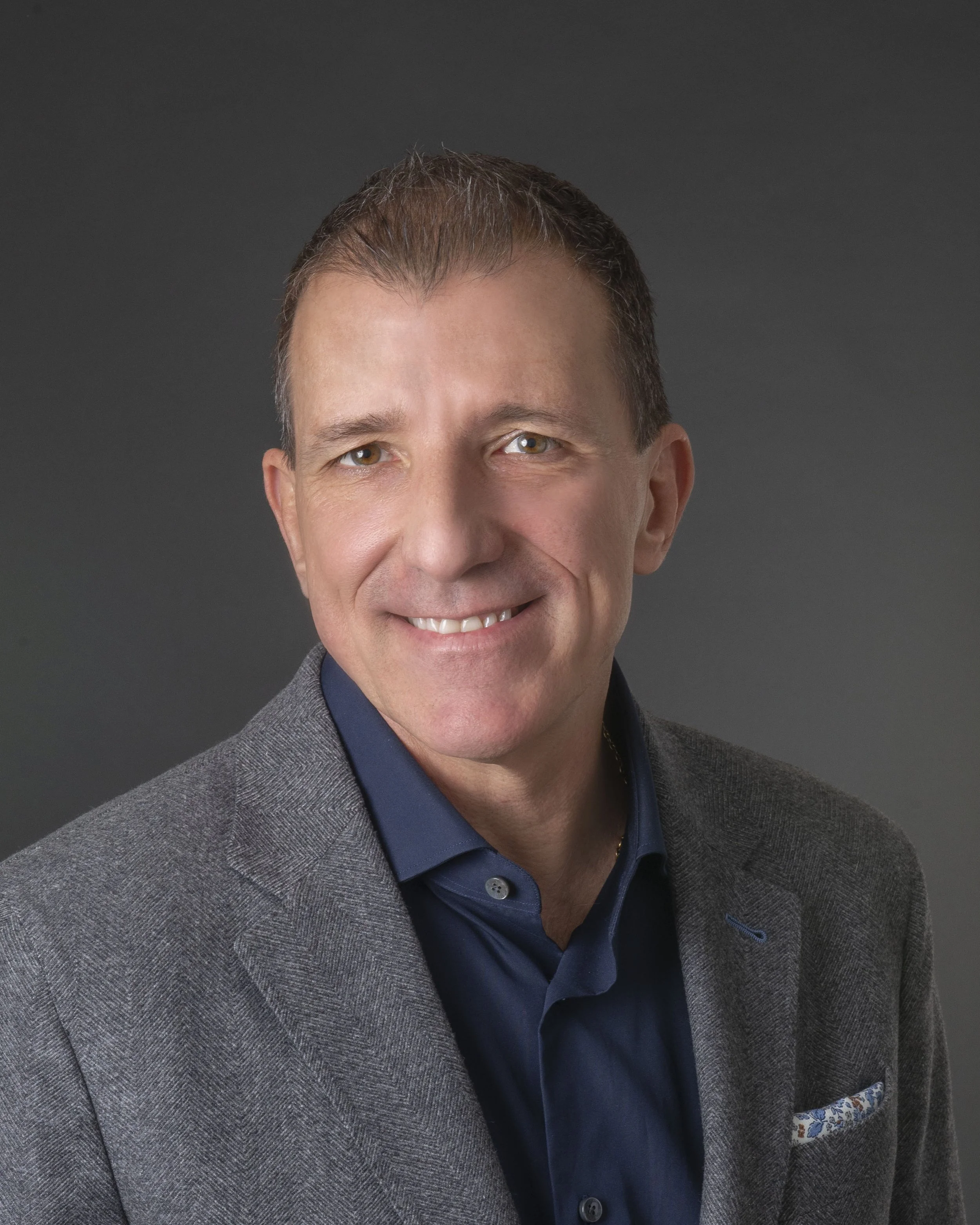 Close-up of a middle-aged man with short brown hair, brown eyes, smiling, wearing a blue shirt, gray blazer, and a patterned pocket square, against a dark gray background.