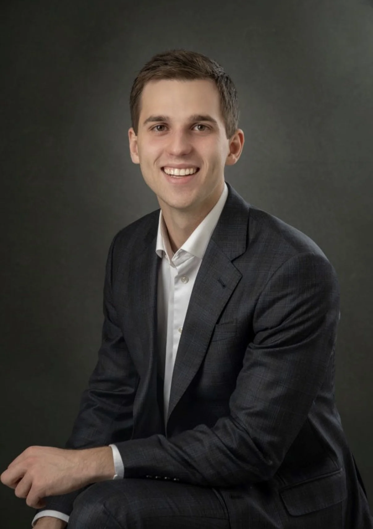 A young man with brown hair wearing a dark gray plaid suit and white shirt, sitting and smiling against a dark background.