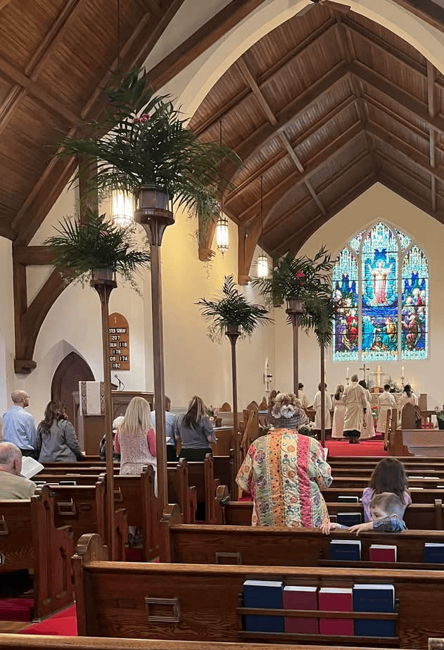 Inside a church with wooden pews, a high arched ceiling, tall lamps with greenery, a colorful stained glass window, and people attending a service.