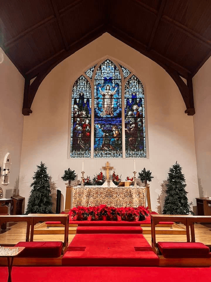 Inside a church with a high vaulted wooden ceiling, a stained glass window depicting a religious scene, an altar with a gold cross, two candles, Christmas decorations including poinsettias and small decorative trees, and red carpeting.